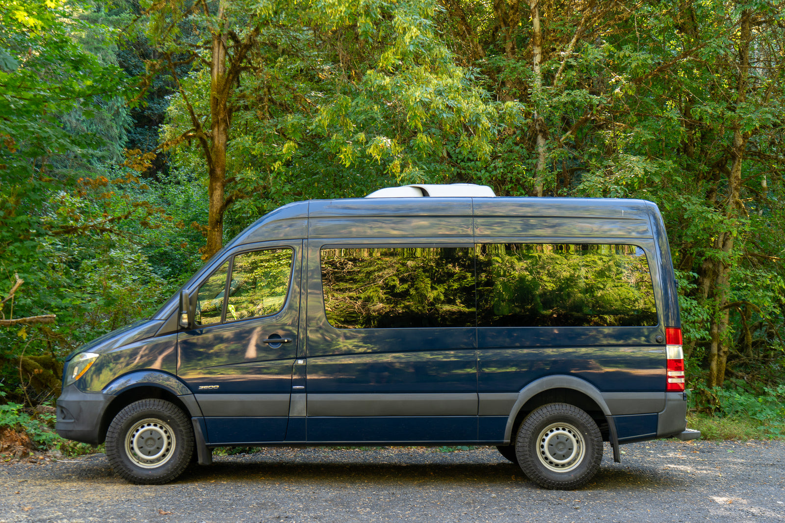 Dark blue Mercedes Sprinter RV rental parked in a lush green forest, ready for outdoor adventure.