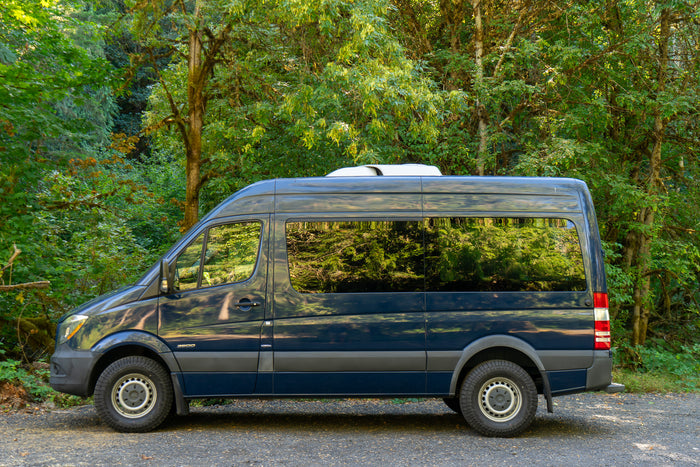 Dark blue Mercedes Sprinter RV rental parked in a lush green forest, ready for outdoor adventure.