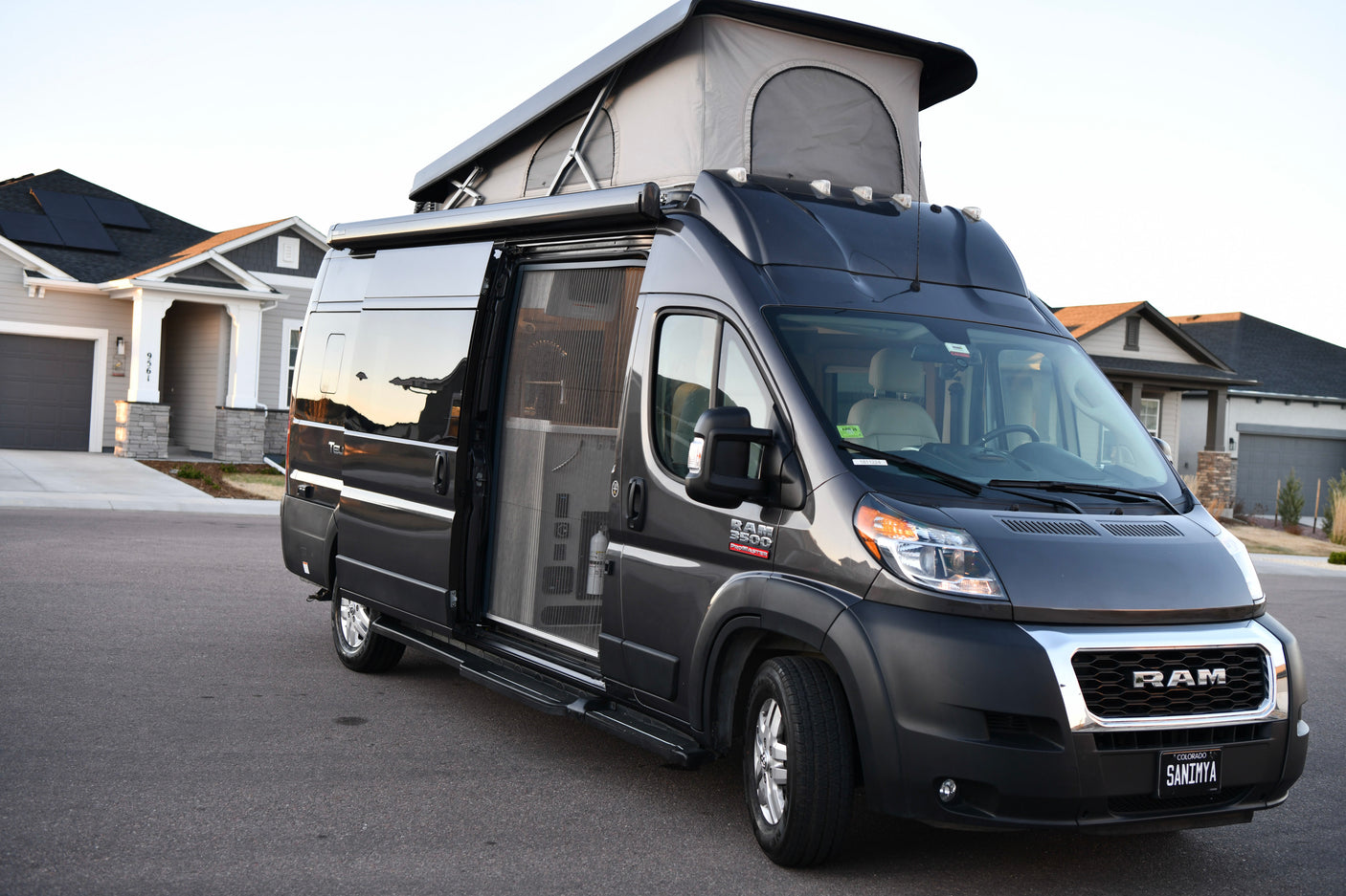 This dark grey pop-top Ram ProMaster camper van RV rental with awning & open side door is parked by a house.