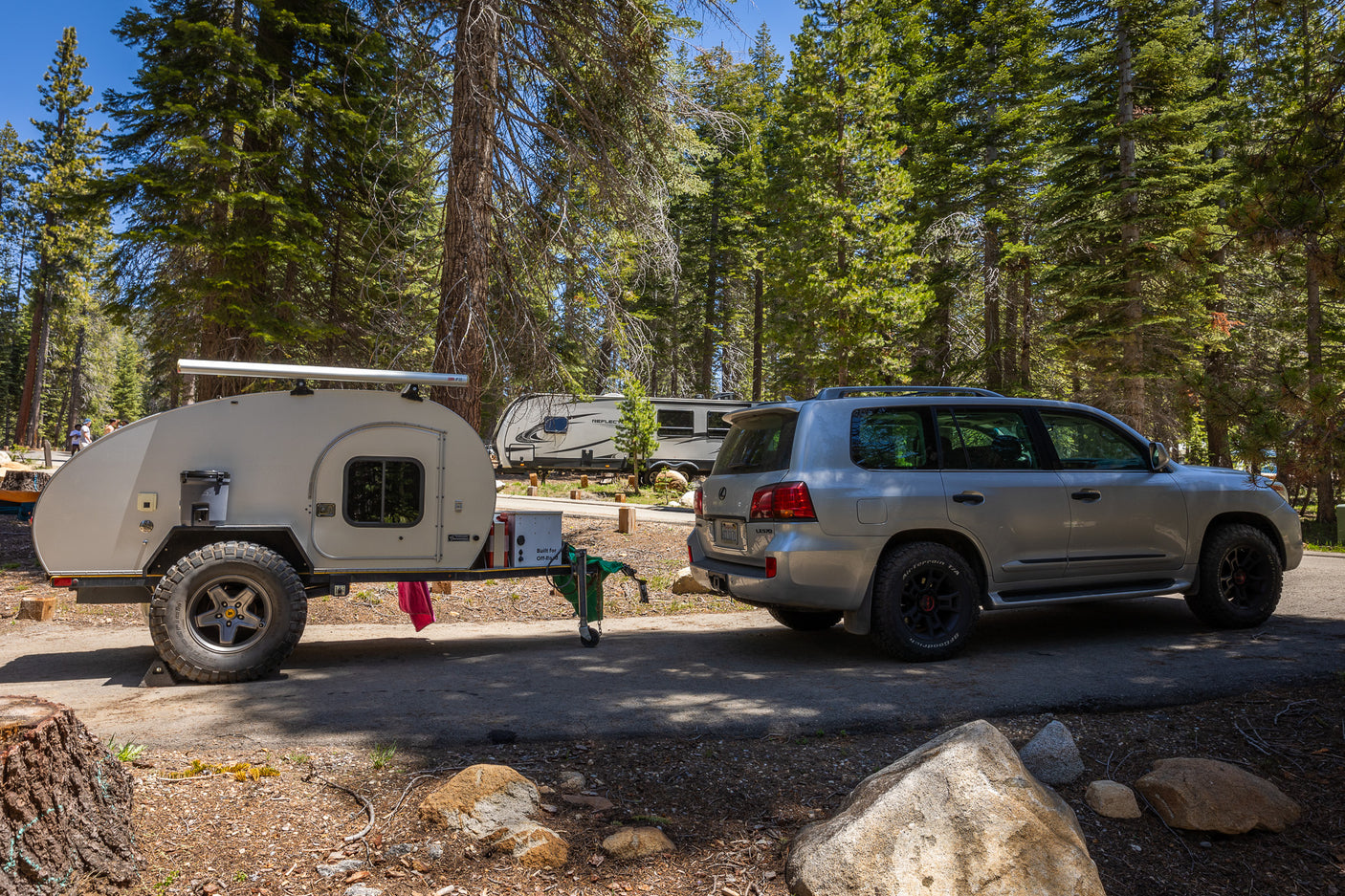 A silver SUV towing a beige teardrop trailer at a sunny forest campsite, perfect for an RV rental trip.