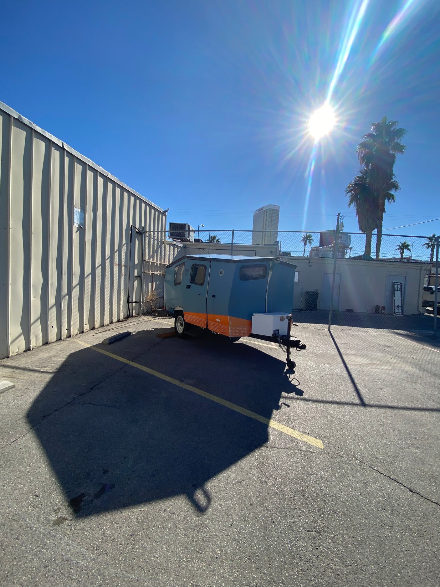 Sunny view of a blue and orange compact rv rental parked in an asphalt lot next to a metal building, with sun glare.