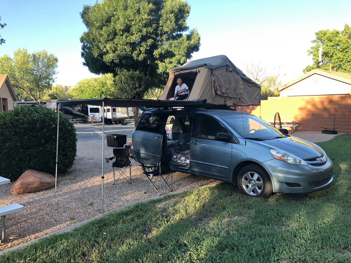 and ladder; a compact alternative to an rv rental for camping.|Cozy rv rental interior: A comfortable bed with a view of a cabin from inside a rooftop tent.|Minivan with rooftop tent and awning set up like an RV rental. Child smiles from tent in sunny campground.|Inside a minivan — photo 7