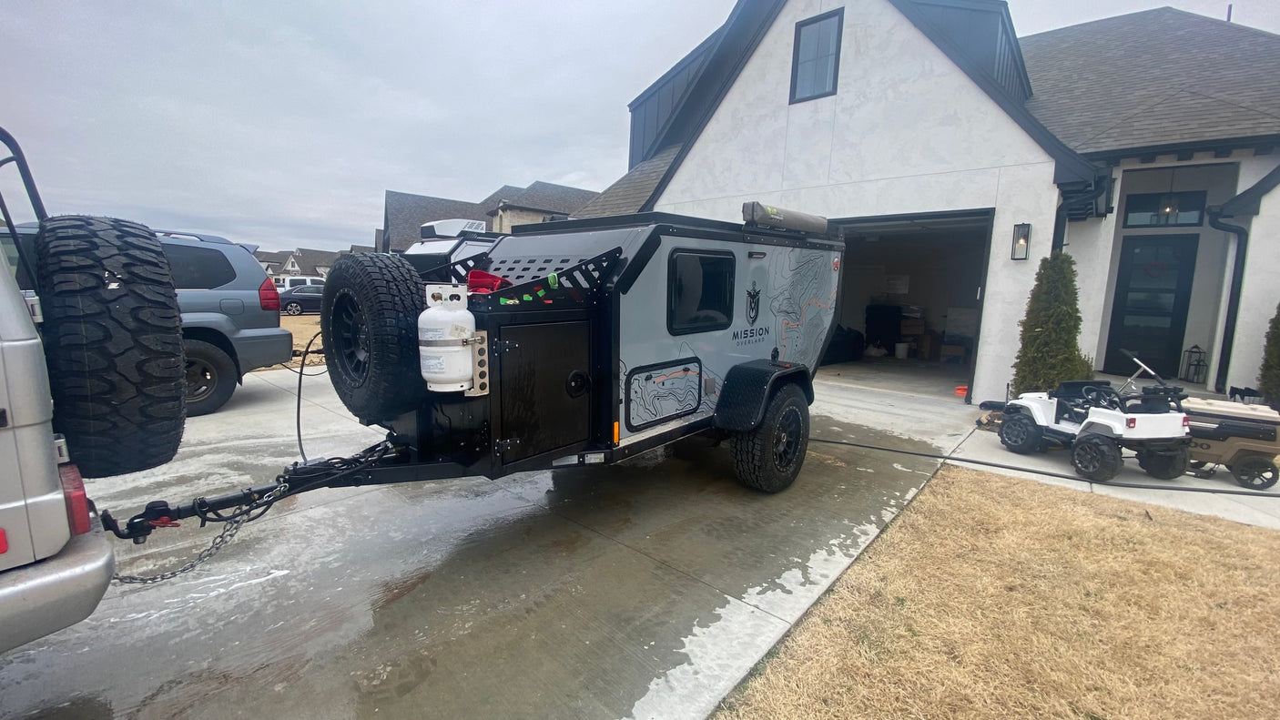 Mission Overland trailer prepped on driveway, perfect for an 'rv rental' outdoor adventure. Note propane tanks & spare tires.