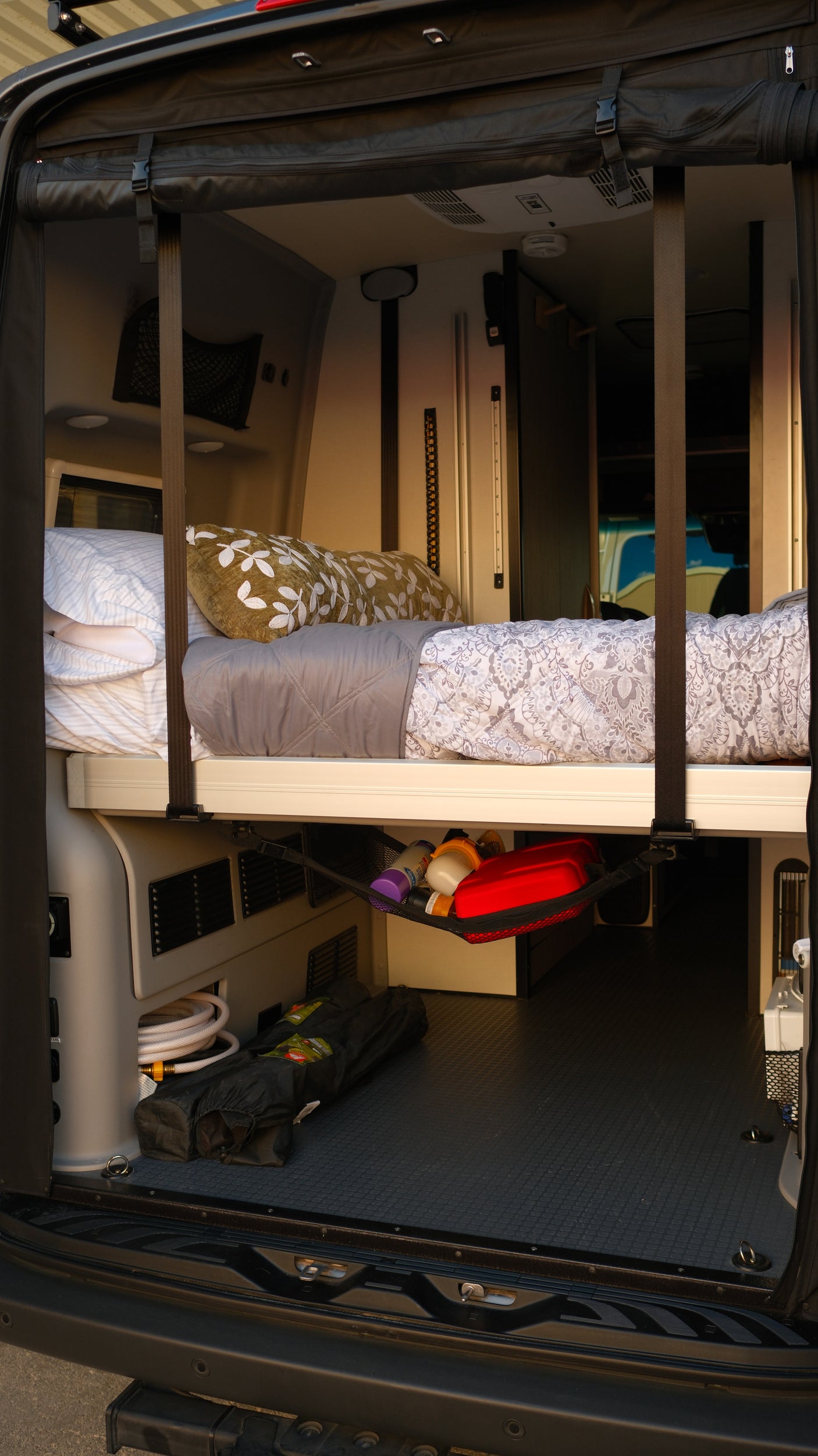 RV rental interior: elevated bed with bedding, under-bed storage with a hose and mesh net, ideal for camper van trips.