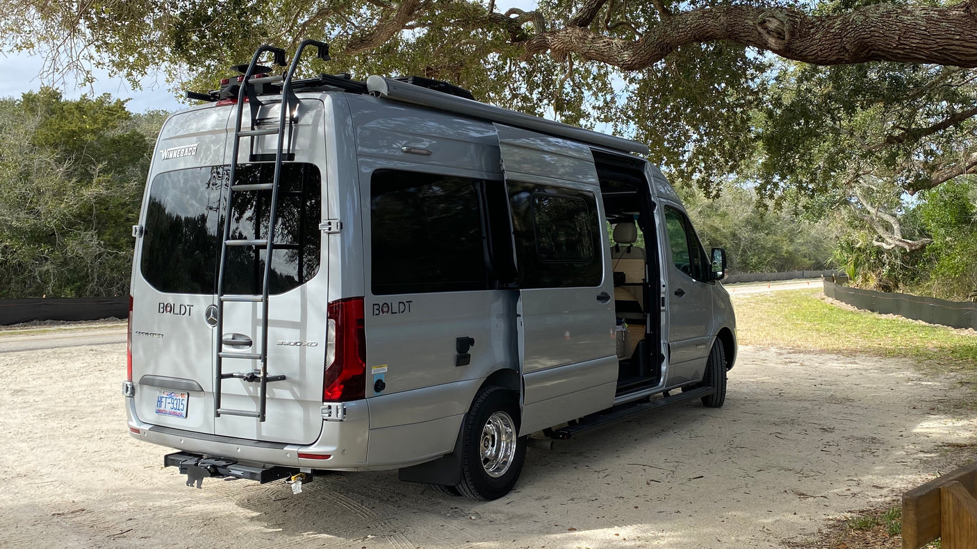 Gray Winnebago Boldt camper van, ideal for rv rental, parked on sand under an oak tree. Its side door is open, showing the interior.
