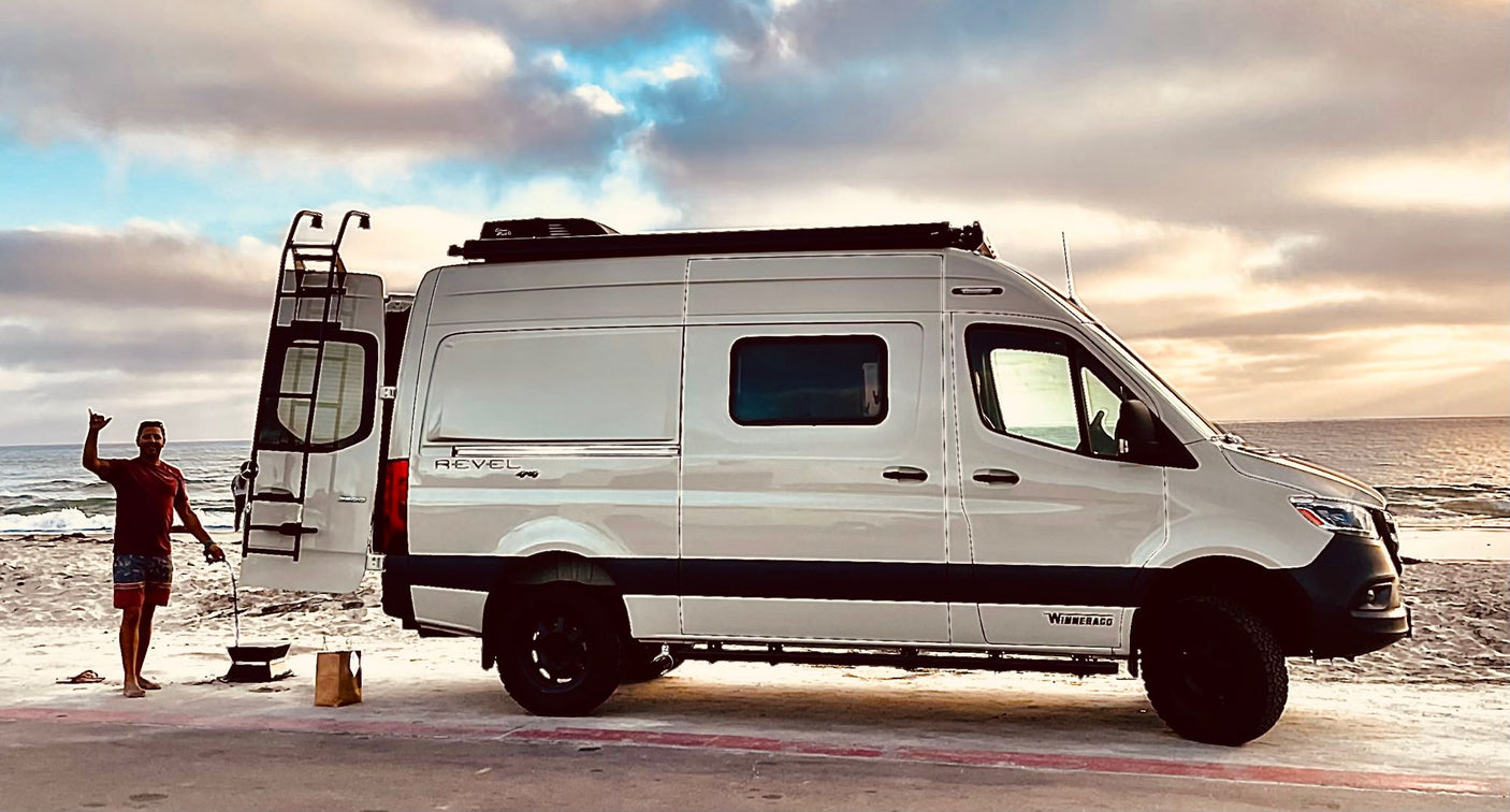 Enjoying the beach with an RV rental. A man stands beside a white Winnebago Revel camper van by the ocean at sunset.