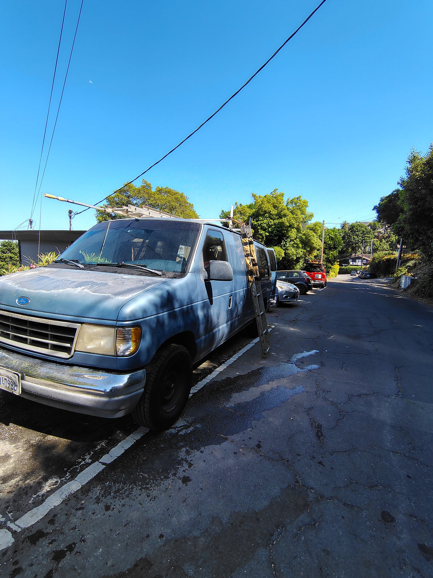 Blue Ford van with a ladder, on a sunlit street. This model is often used for RV rental conversions. A crescent moon is visible.