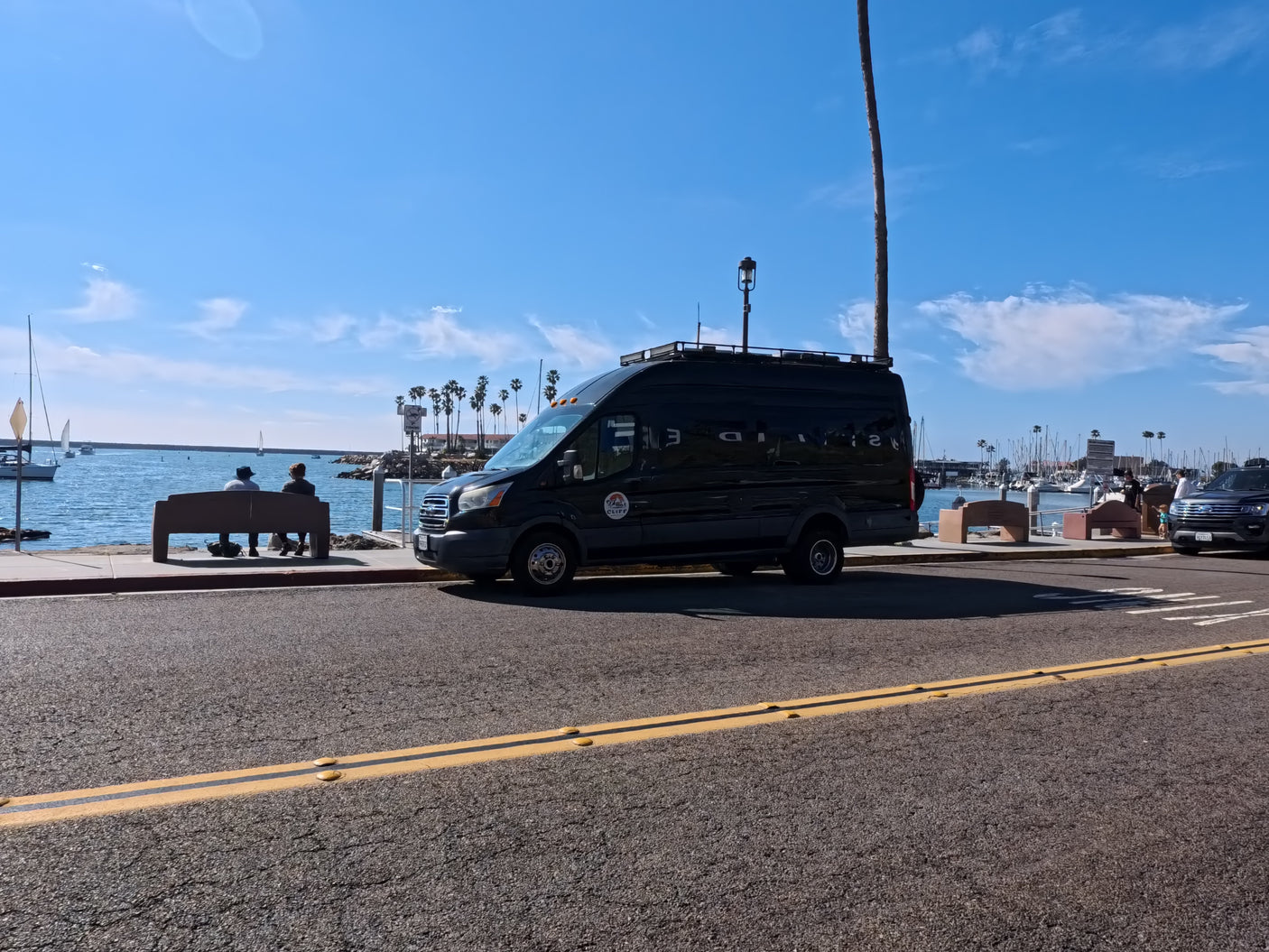 Black RV rental van beside a sunny marina, boats, and palm trees. Perfect for coastal adventures.