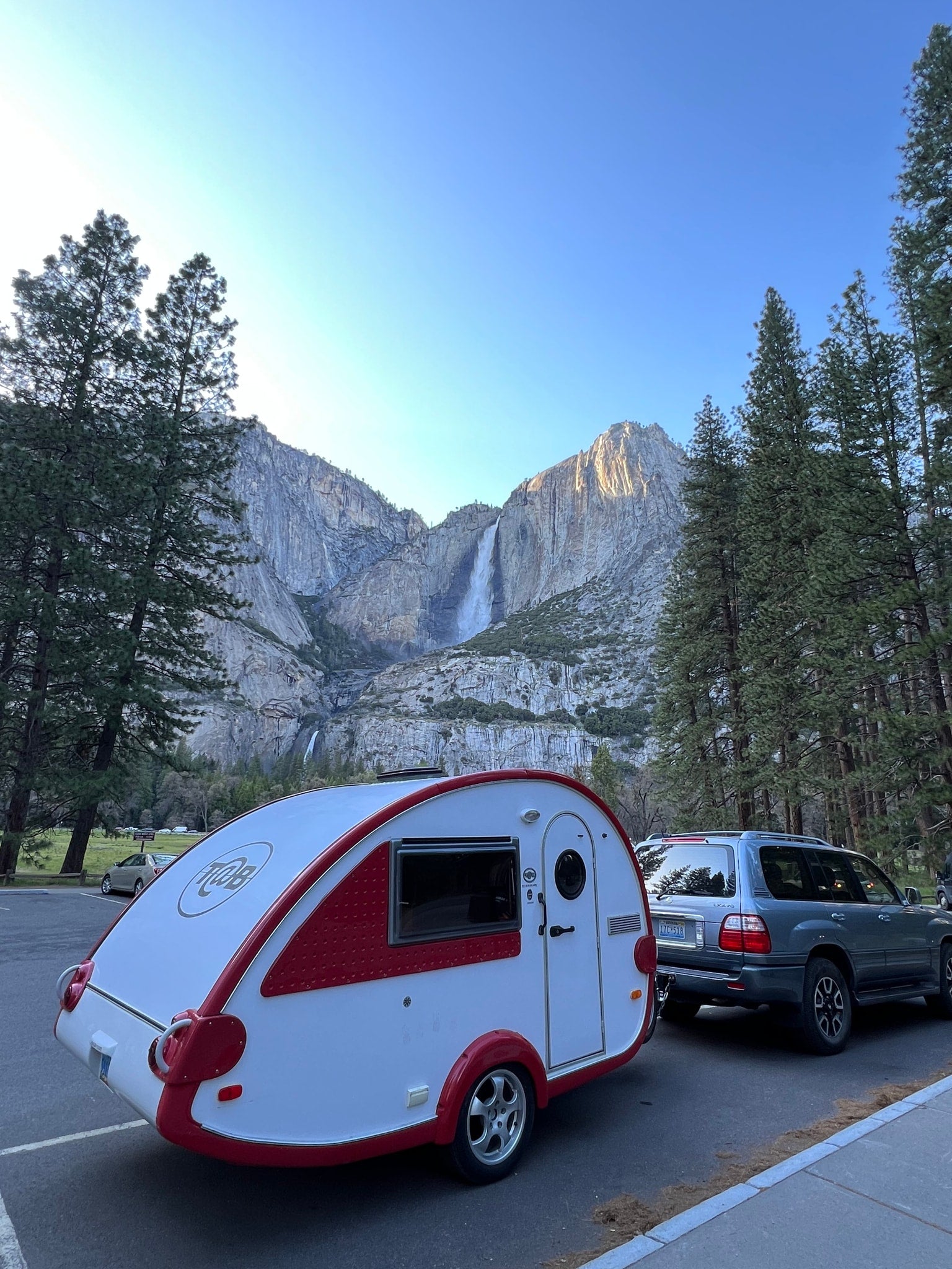 amidst vibrant blue wildflowers and pine trees.|A compact white and red T@B teardrop RV rental parked on a wooden deck by a tranquil river and lush trees.|Scenic Yosemite adventure with an RV rental! A teardrop camper and SUV are parked with Yosemite Falls in the background.|A silver SUV tows a white and red T@B travel trailer (like an rv rental) on a winding road through a peaceful
