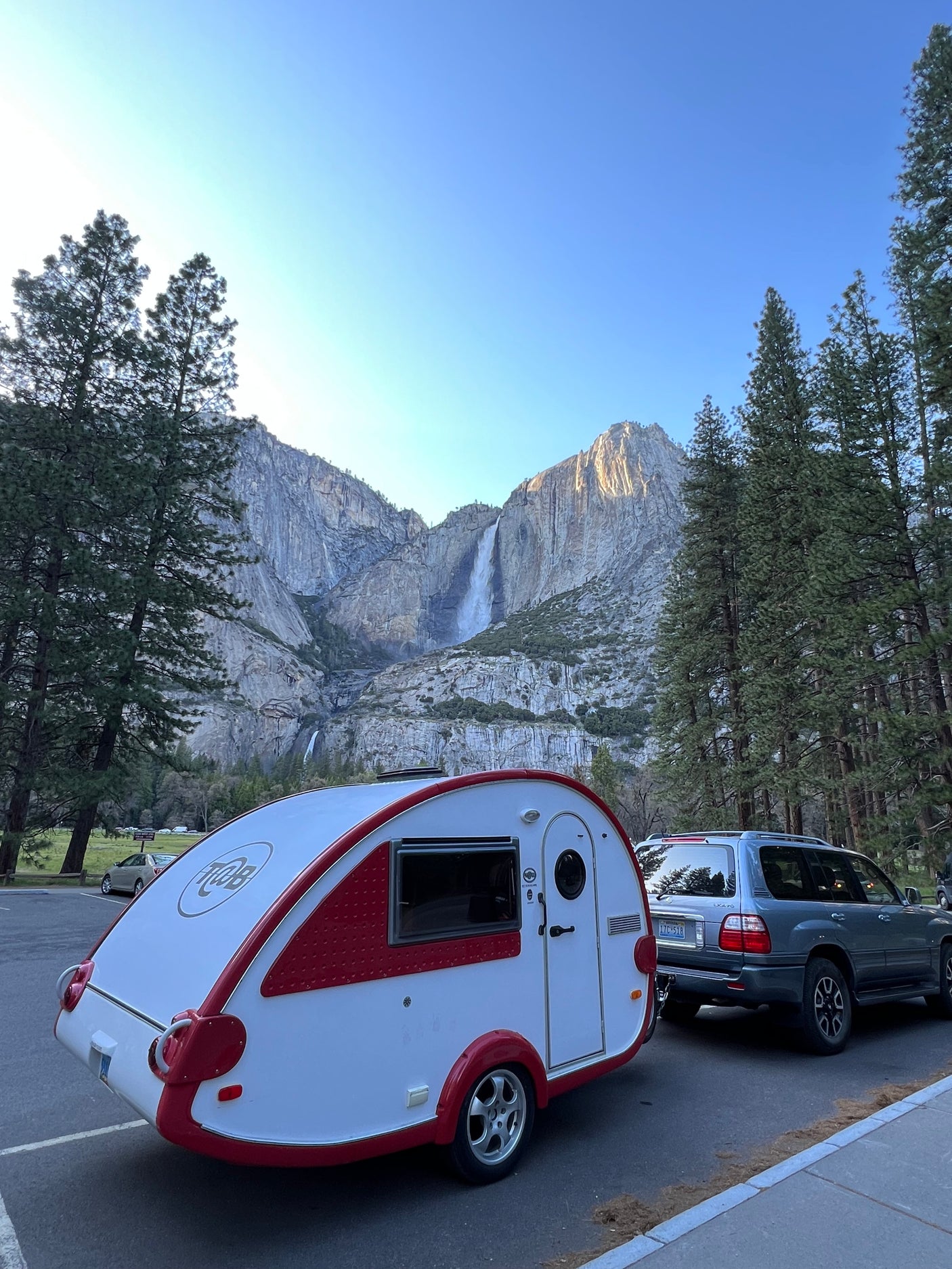 amidst vibrant blue wildflowers and pine trees.|A compact white and red T@B teardrop RV rental parked on a wooden deck by a tranquil river and lush trees.|Scenic Yosemite adventure with an RV rental! A teardrop camper and SUV are parked with Yosemite Falls in the background.|A silver SUV tows a white and red T@B travel trailer (like an rv rental) on a winding road through a peaceful