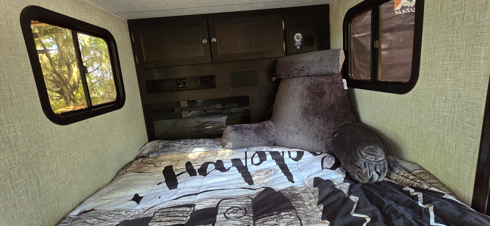 Cozy RV rental bedroom featuring a large bed with a backrest pillow, dark cabinetry, and bright windows showing lush trees outside.