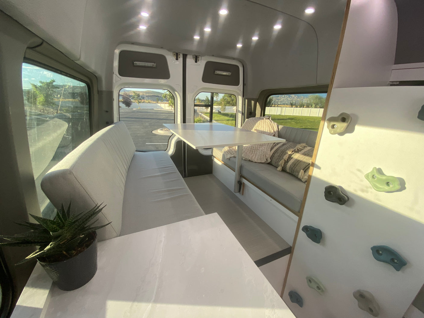 Bright RV rental interior featuring a dinette with light gray cushions, a white table, ample windows, and a playful climbing wall.