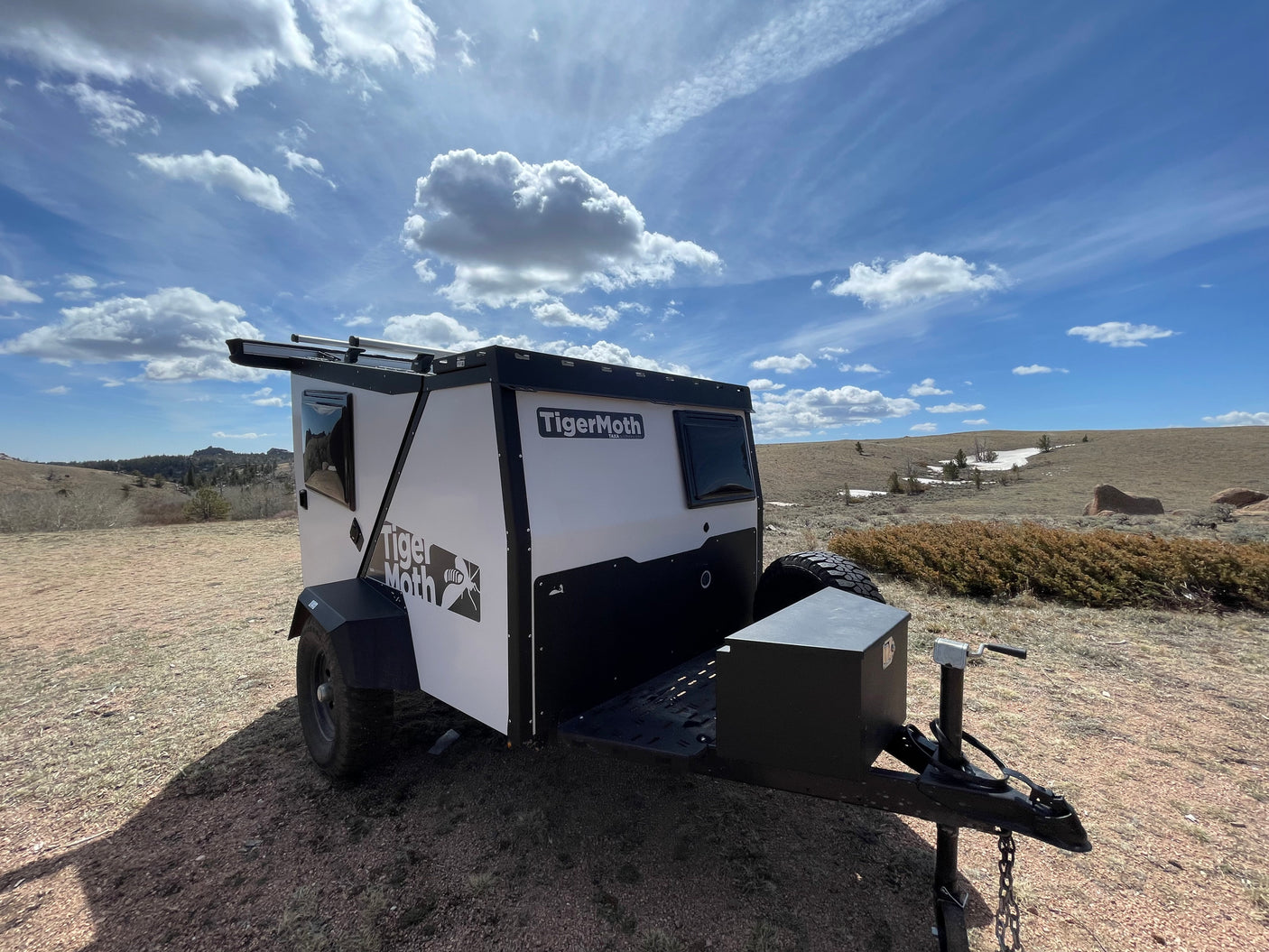 White & black TigerMoth compact RV rental parked on a dirt hill with rocky terrain, under a partly cloudy sky.