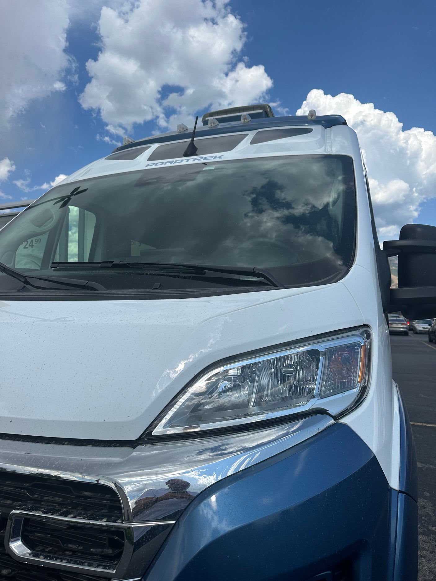 Front view of a white and blue Roadtrek campervan for rv rental. Clouds reflect on the windshield, parked under a blue sky.