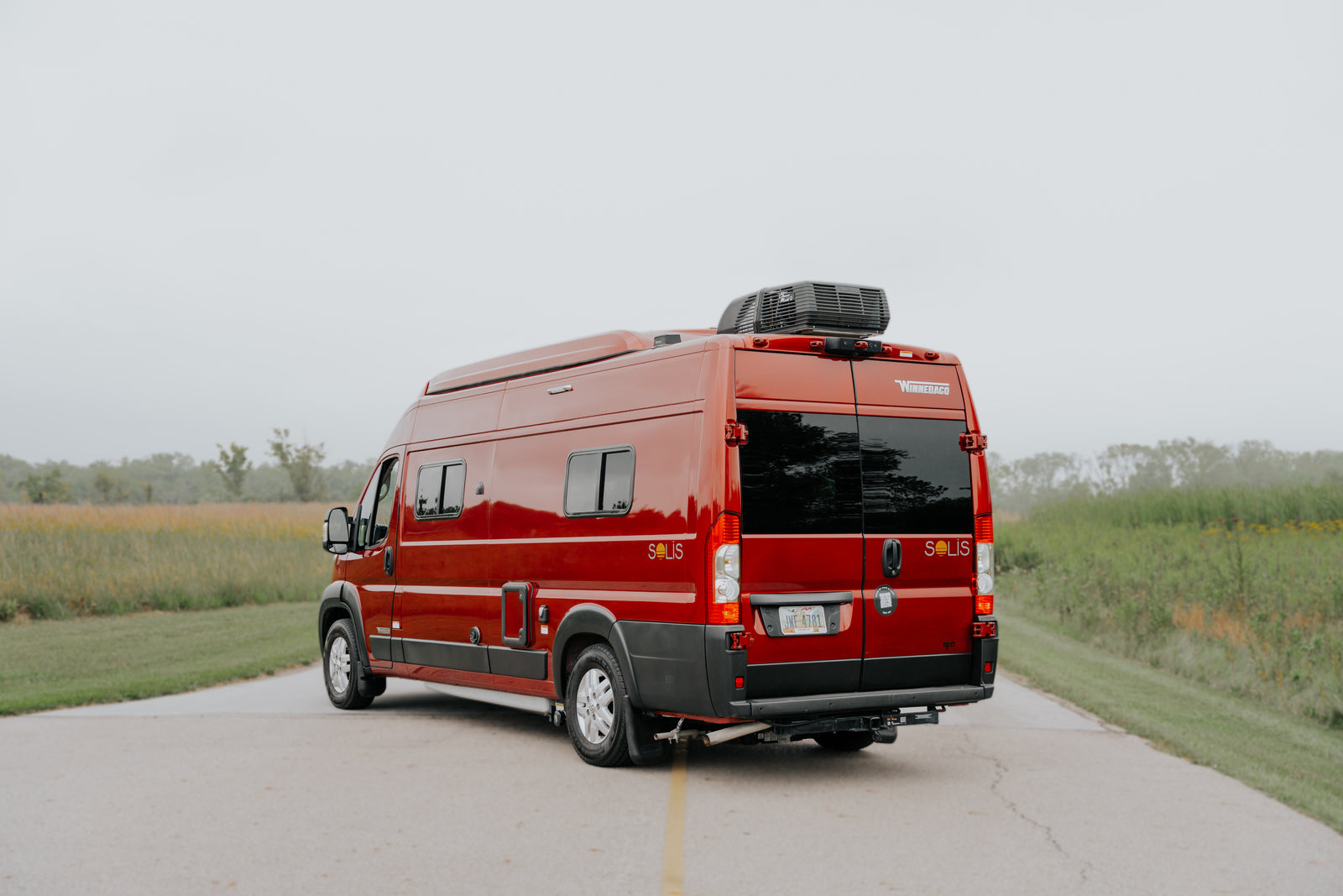A bright red Winnebago Solis camper van, perfect for RV rental, rests on a road, flanked by green fields under an overcast sky.