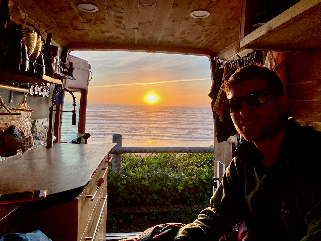 Unwind with an rv rental! Man enjoys a stunning ocean sunset view from his cozy, well-appointed camper van kitchen.