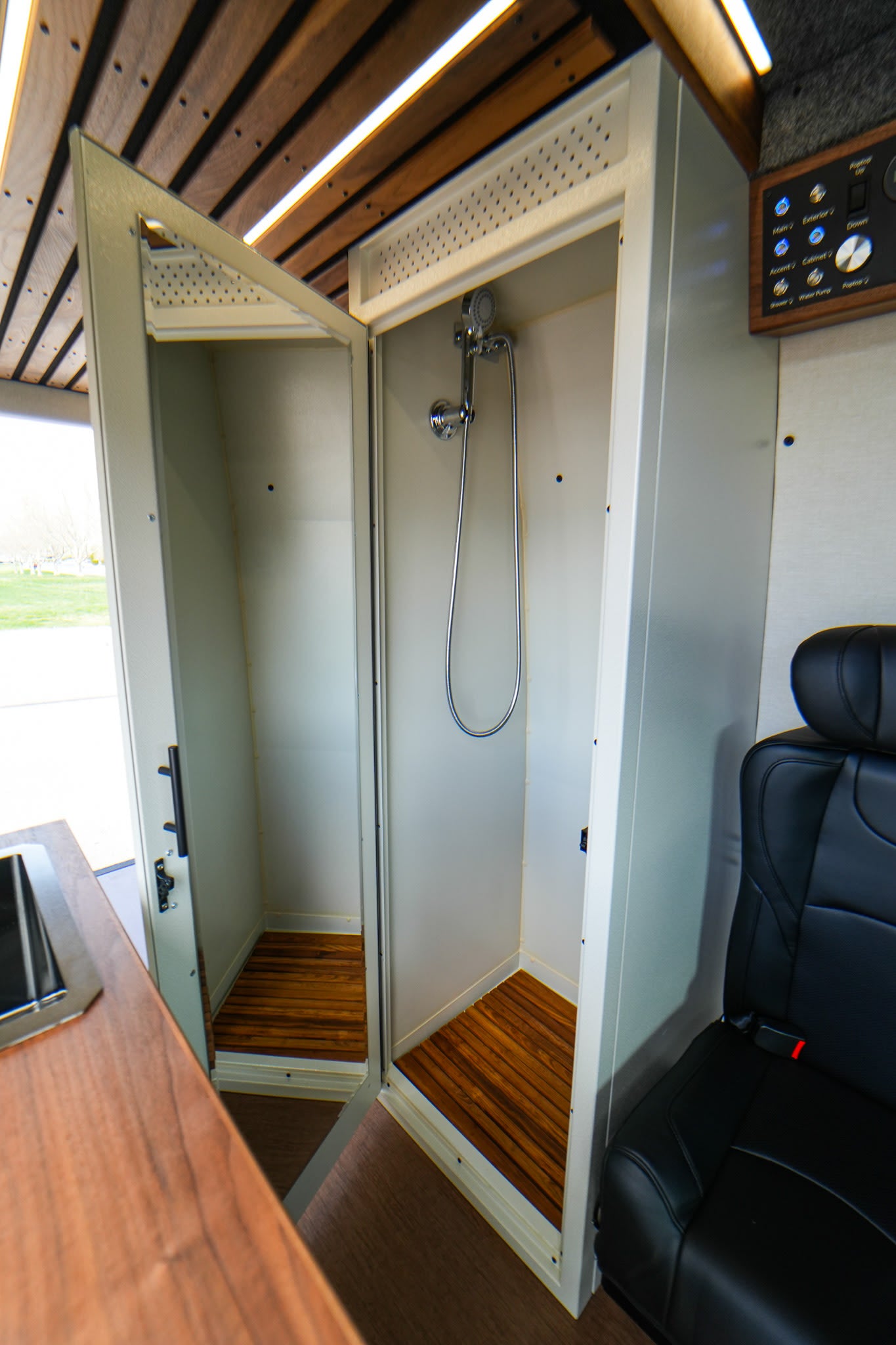 Inside an RV rental: a compact, open shower stall with wood slats, controls & a black leather seat visible. Efficient use of space.