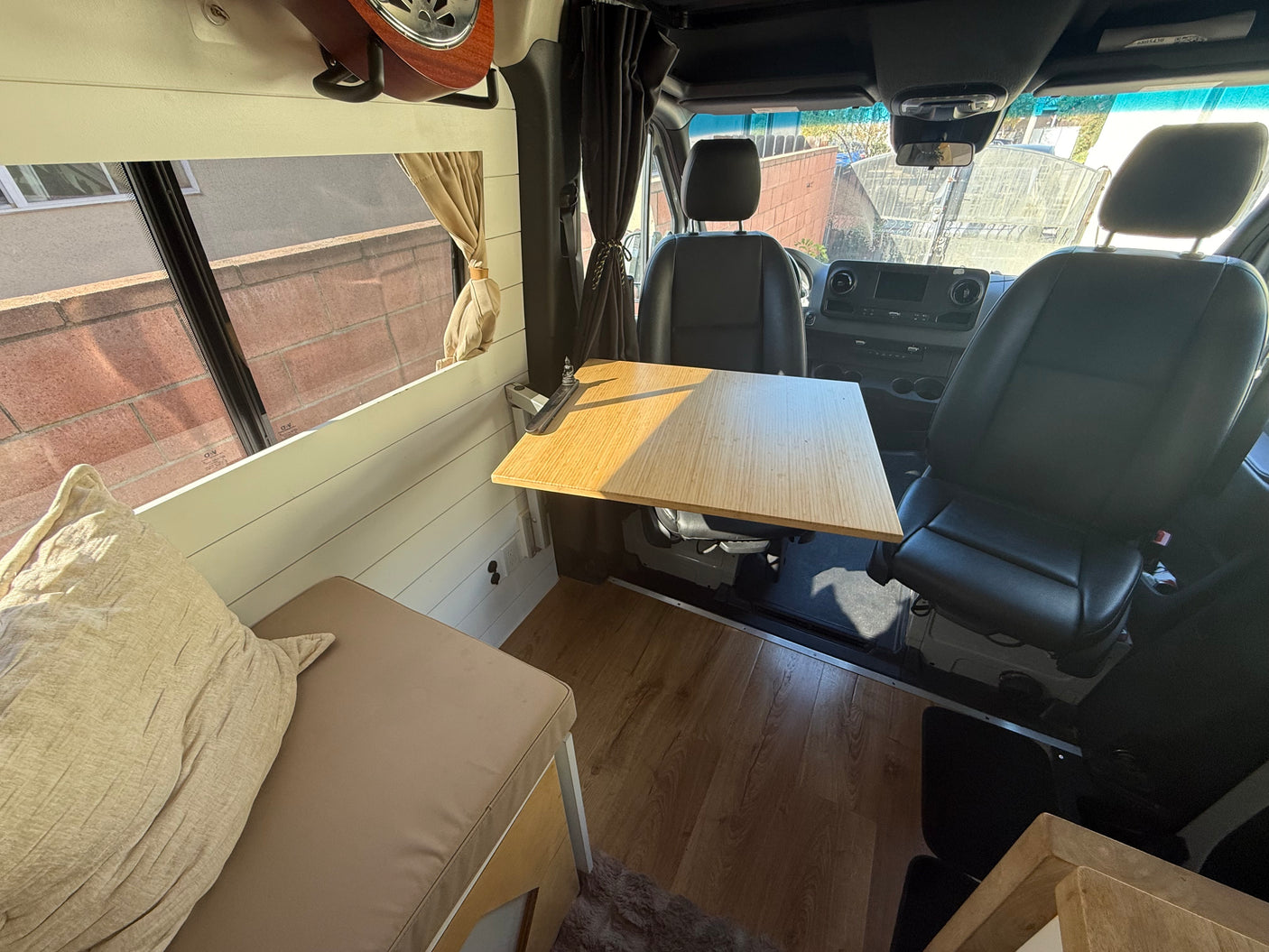 Inside an RV rental camper van: light wood table, tan cushioned bench, driver seats, and dark curtains for comfort.