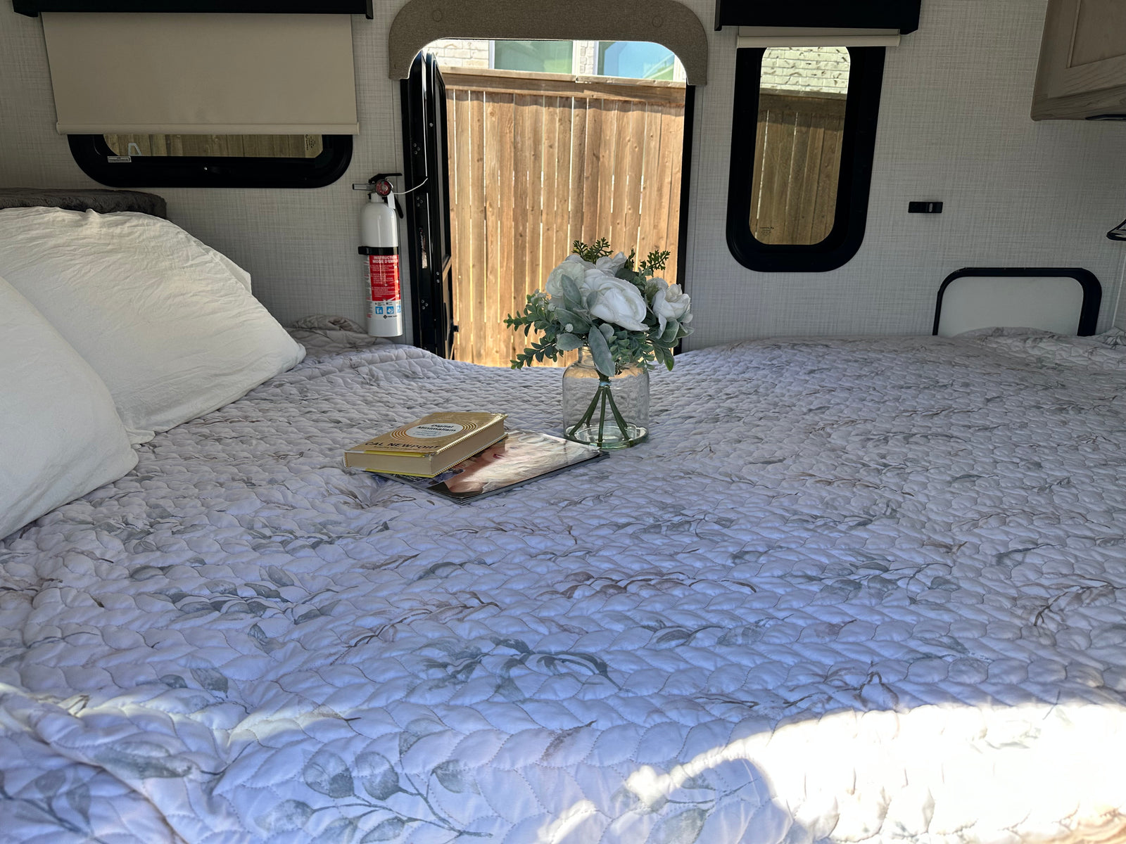 A comfortable bed with a quilted spread, white pillows, books, and flowers inside an RV rental. A fire extinguisher is mounted nearby.