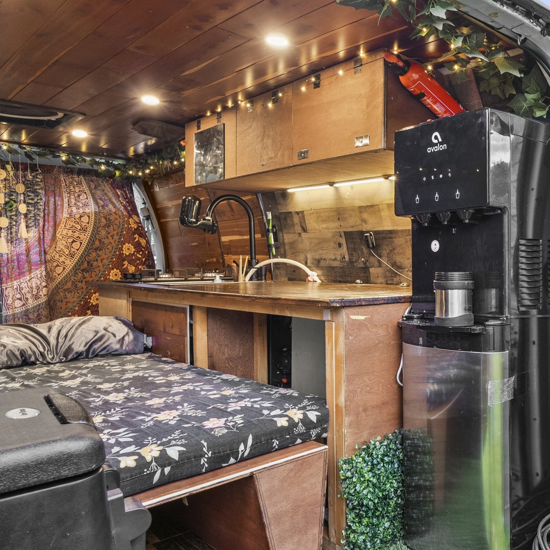 Cozy camper van RV rental interior: a comfy bed with floral mattress, compact wood kitchenette, sink, and water dispenser.