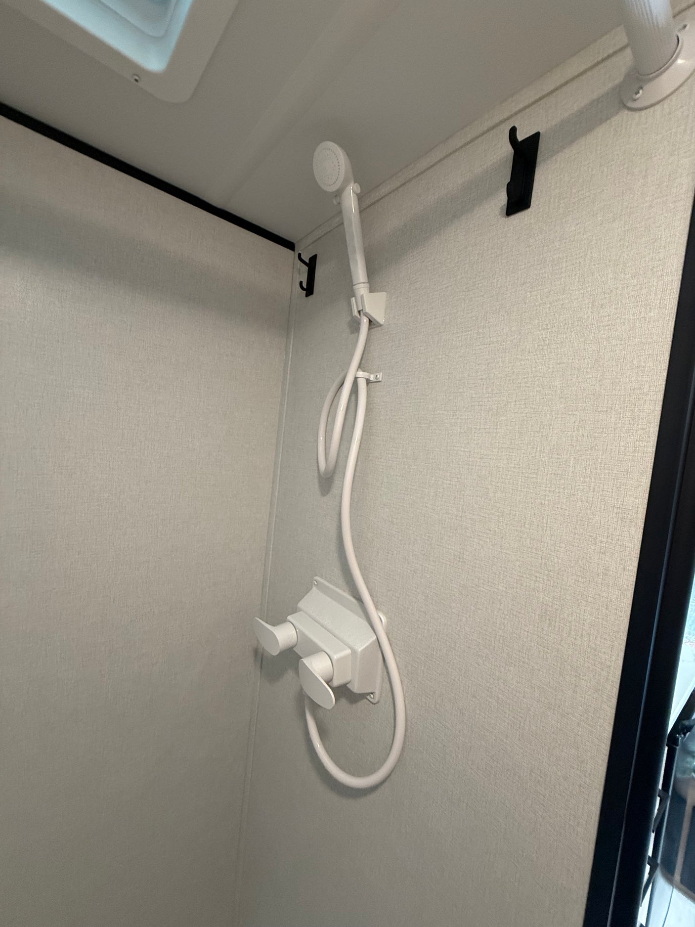 Interior of an RV rental shower featuring a white handheld showerhead, hose, and dual controls on a light textured wall with hooks.