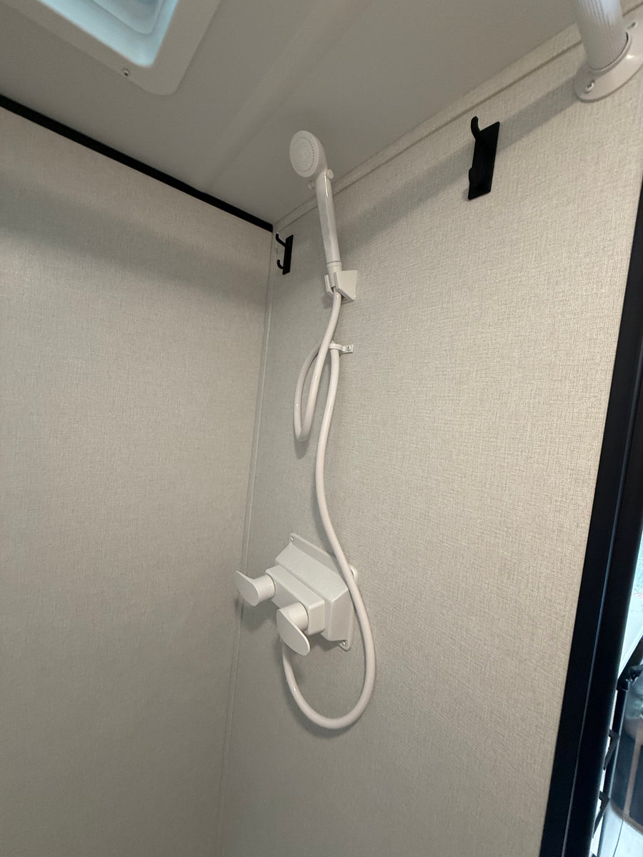 Interior of an RV rental shower featuring a white handheld showerhead, hose, and dual controls on a light textured wall with hooks.