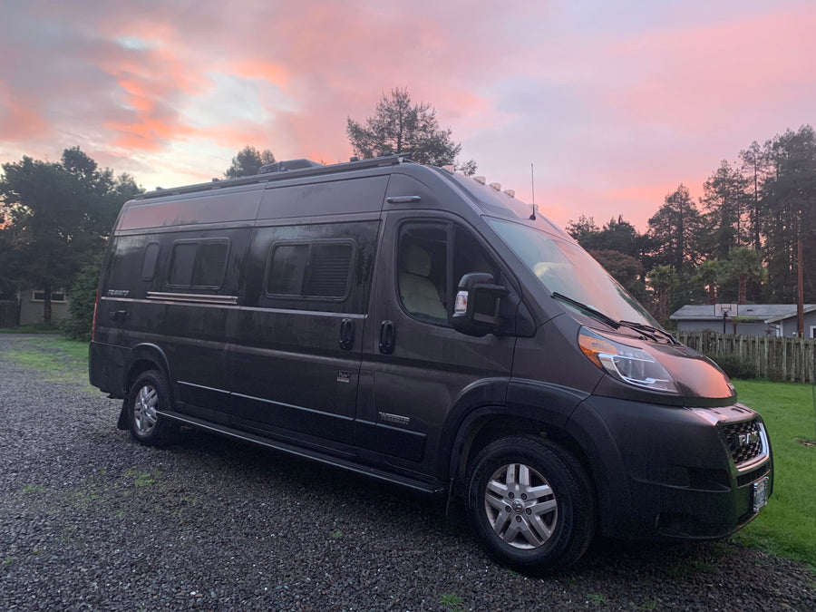 Dark gray Winnebago Travato camper van parked on gravel under a beautiful pink sunset. Ideal for your next rv rental.