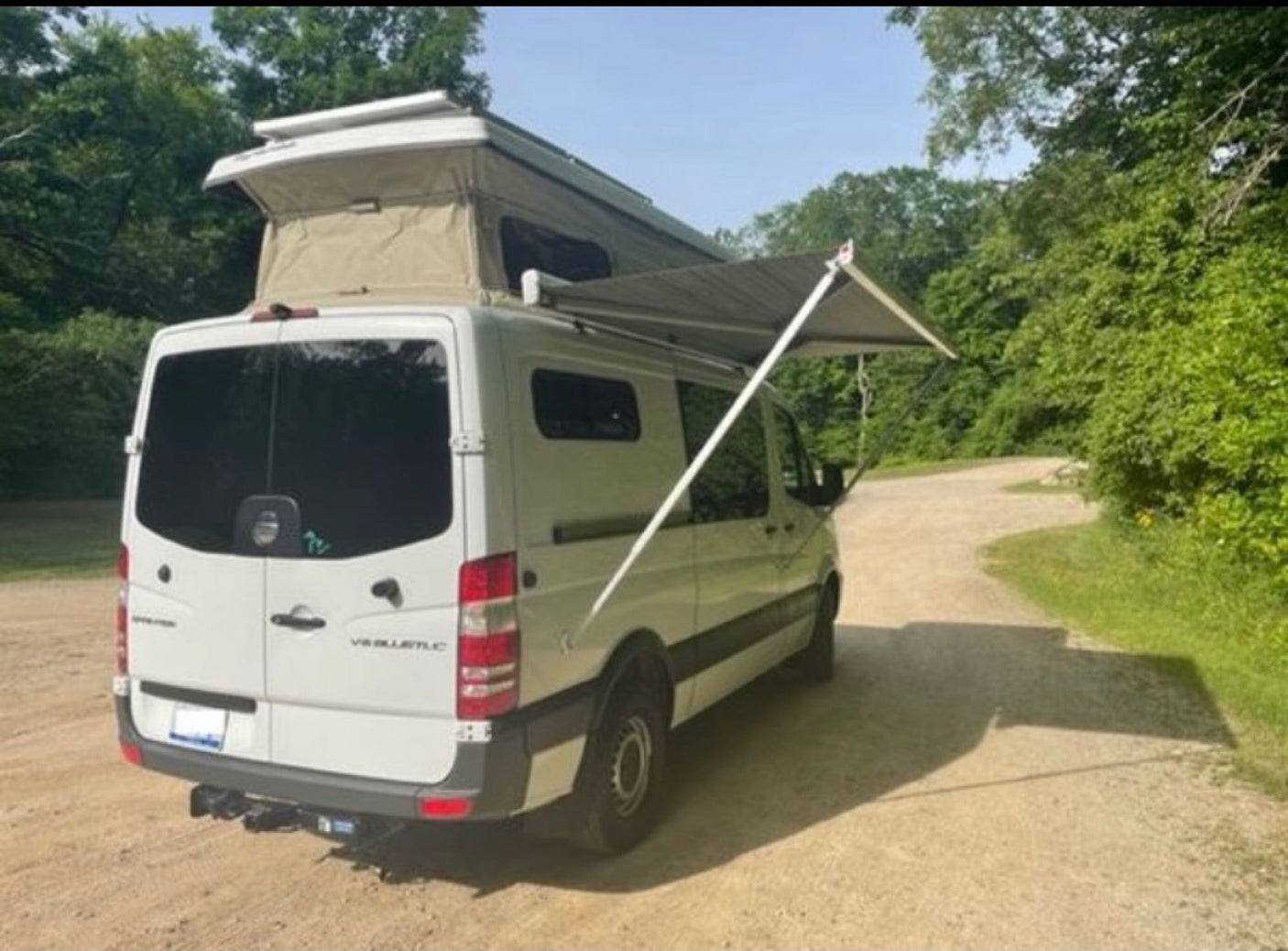 A white Sprinter camper van with a raised pop-top and extended awning on a dirt road. Great rv rental option.|This white Mercedes-Benz Sprinter camper van