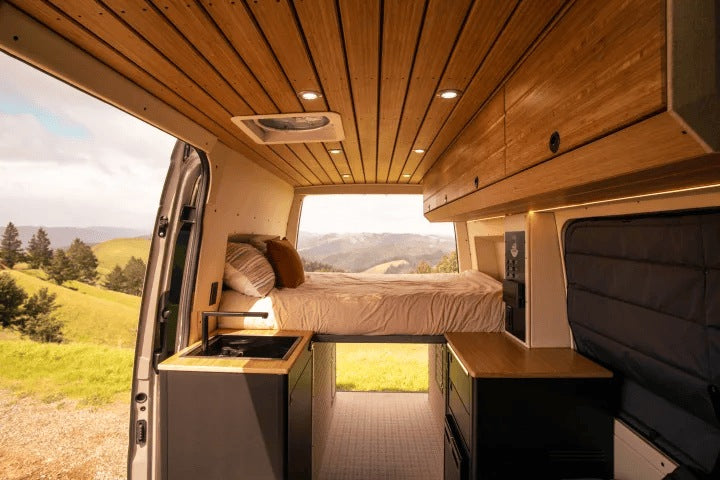 Inside a modern rv rental camper van: wooden ceiling, kitchen sink, and a comfortable bed with scenic mountain views.