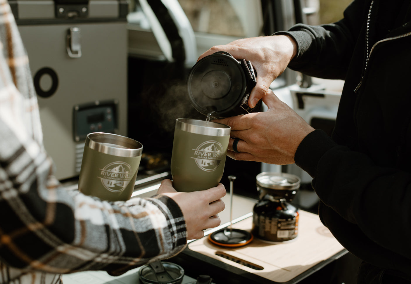 Two people preparing hot drinks in olive green tumblers inside an RV rental, highlighting a cozy camping setup.