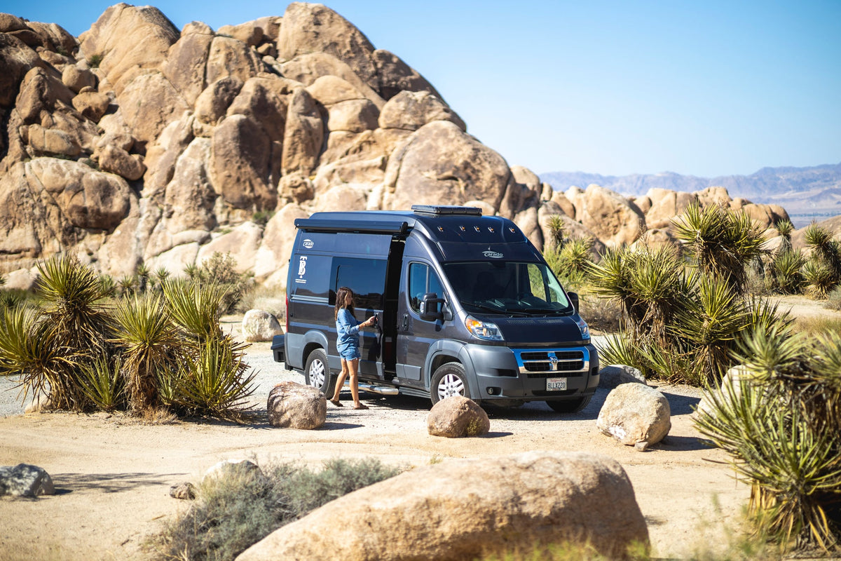 a paddleboard leaning nearby. Perfect for outdoor van life.|An RV rental camper van parked in a sunny desert landscape with large rock formations. A woman exits the side door.|RV rental adventure: Man & dog from pop-top Carado van. Amber lights glow under a cloudy sky with saguaros.|Man loads gear into a storage box on the back of an RV rental van amidst Joshua trees in a sunny desert.|Explore the comfort of an RV rental: compact bathroom with shower — photo 4