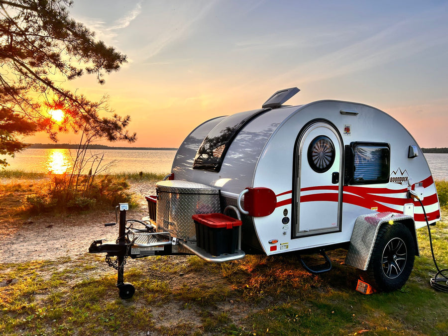 Enjoy an RV rental adventure! A white teardrop camper with red accents sits by a lake at sunset