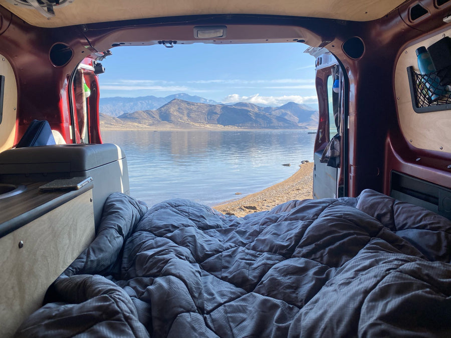 surrounded by towering red rock mountains.|RV rental: Person enjoys a tranquil lake & mountain view from inside a camper van.|Relax in this RV rental! A cozy bed overlooks a stunning lake and mountains from inside the van.|Red Ram ProMaster City van