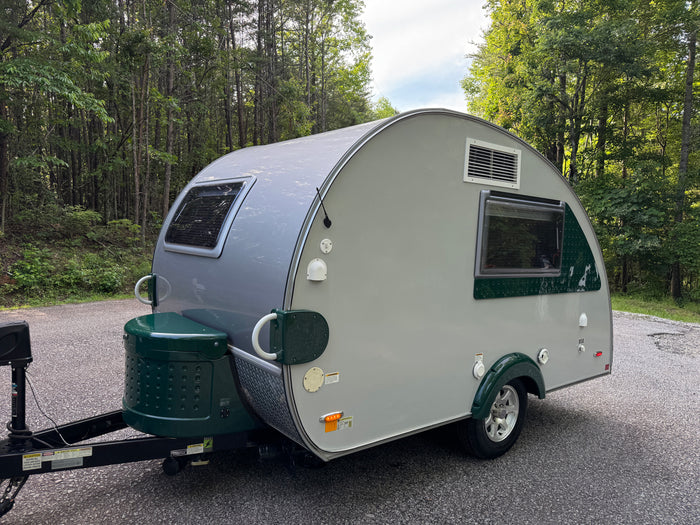 Grey & green teardrop RV rental parked on an asphalt road in a green forest, ready for travel.