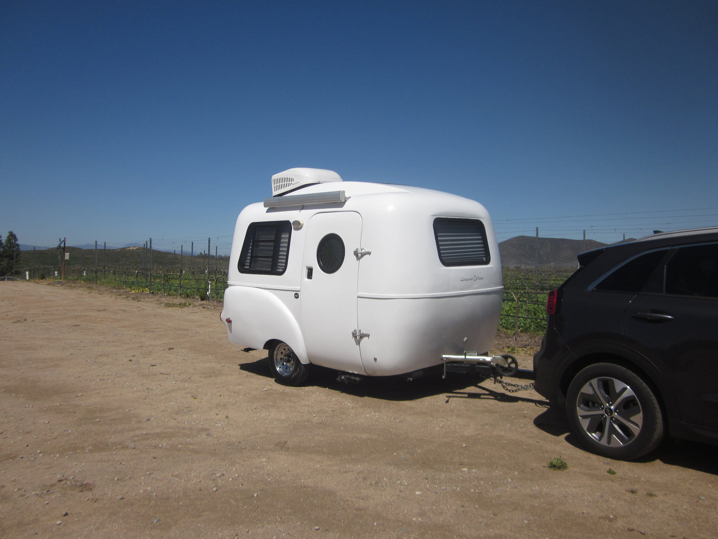 A white Happier Camper travel trailer, ideal for RV rental, hitched to an SUV in a sunny vineyard setting.