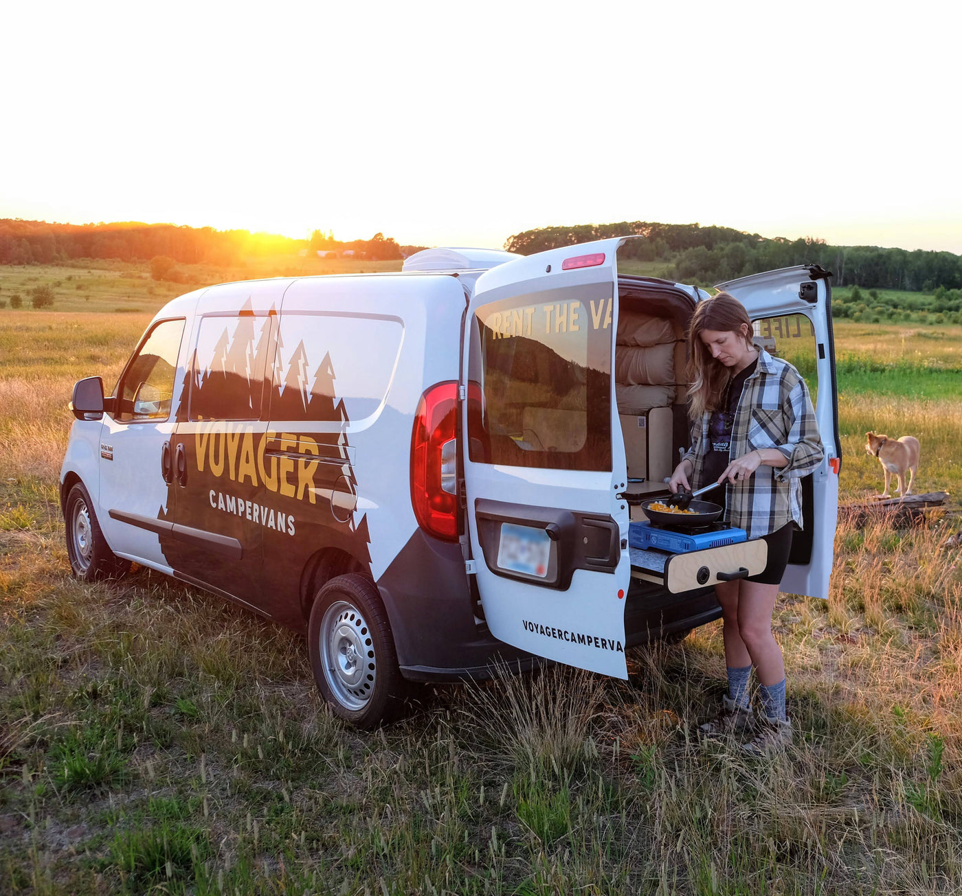 Woman cooks a meal at sunset from her white Voyager campervan rv rental. Her dog watches in the grassy field. — photo 17