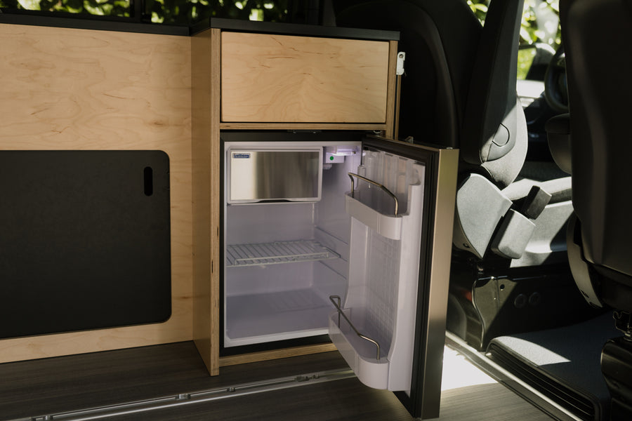 Compact RV rental interior featuring a built-in wooden kitchenette with an open mini-fridge and vehicle seating.