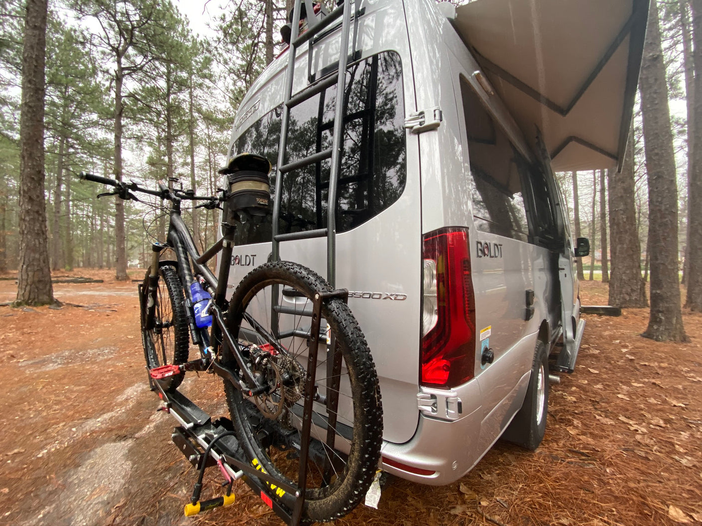 RV rental adventure: Silver camper van with a mountain bike on a rear rack & side awning, parked in a pine forest.