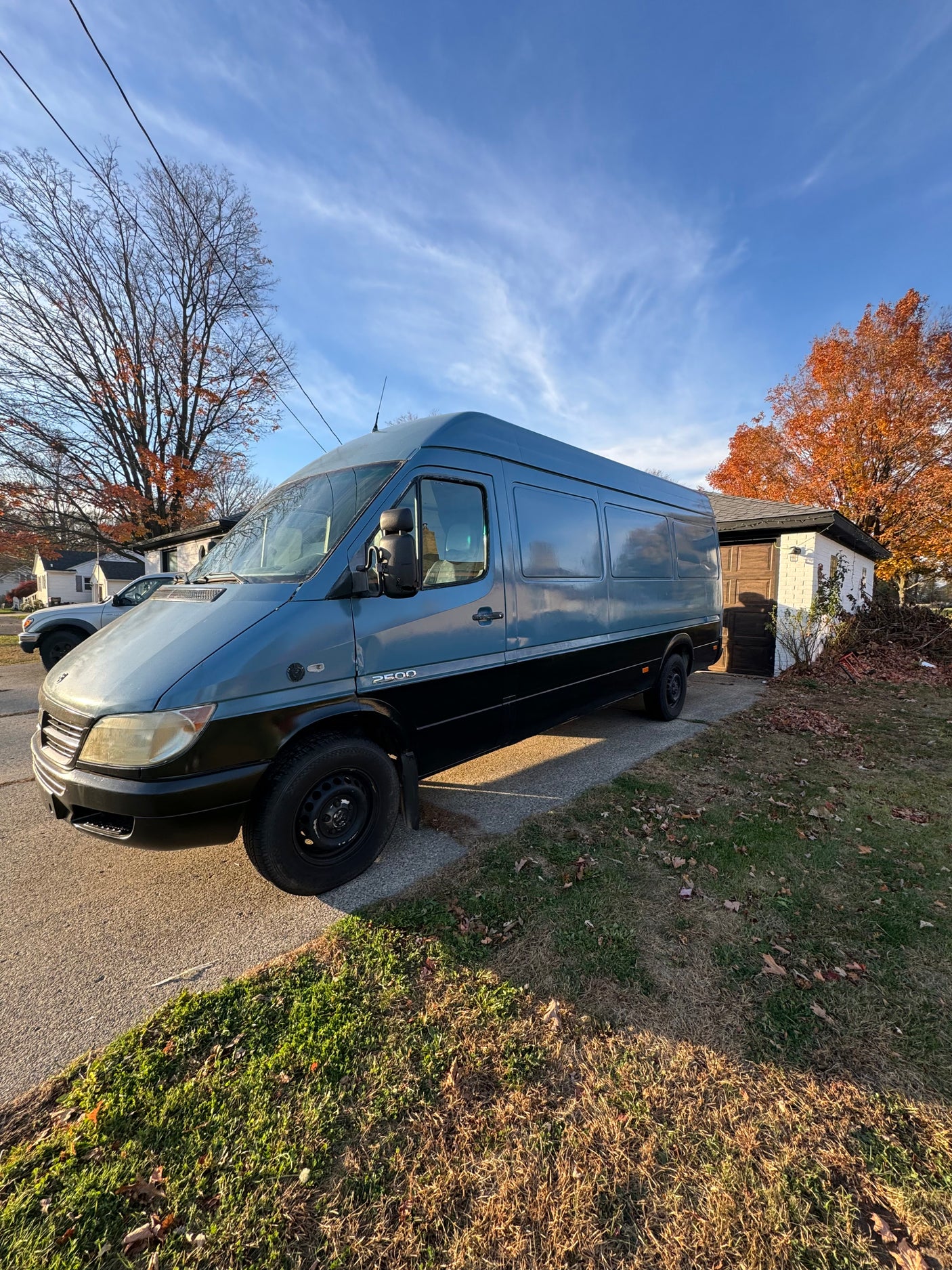 This blue & black Mercedes Sprinter van, suitable for **RV rental**, is parked on a sunlit driveway with autumn foliage.