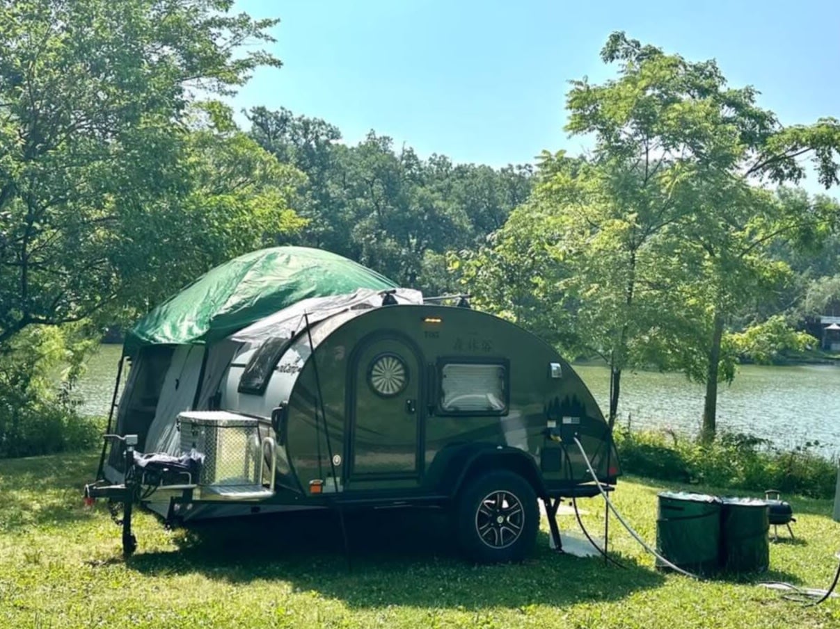 sits by a lake among trees. Barrels & a small grill are visible.|RV rental camping setup with a teardrop trailer's open kitchen — photo 3