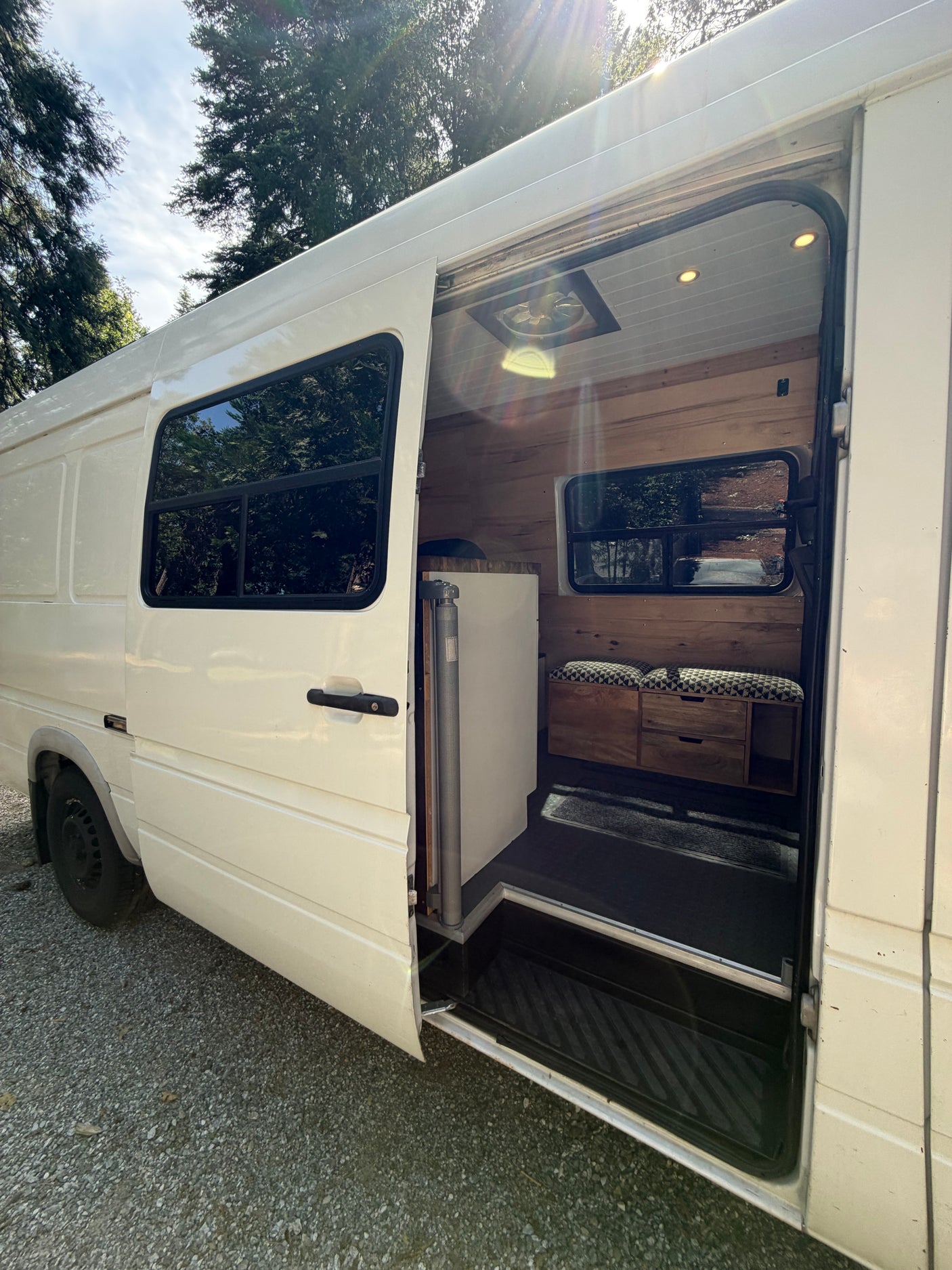 Open side door of a white camper van showing a custom wood interior, bench seat, and drawers. A perfect RV rental choice!
