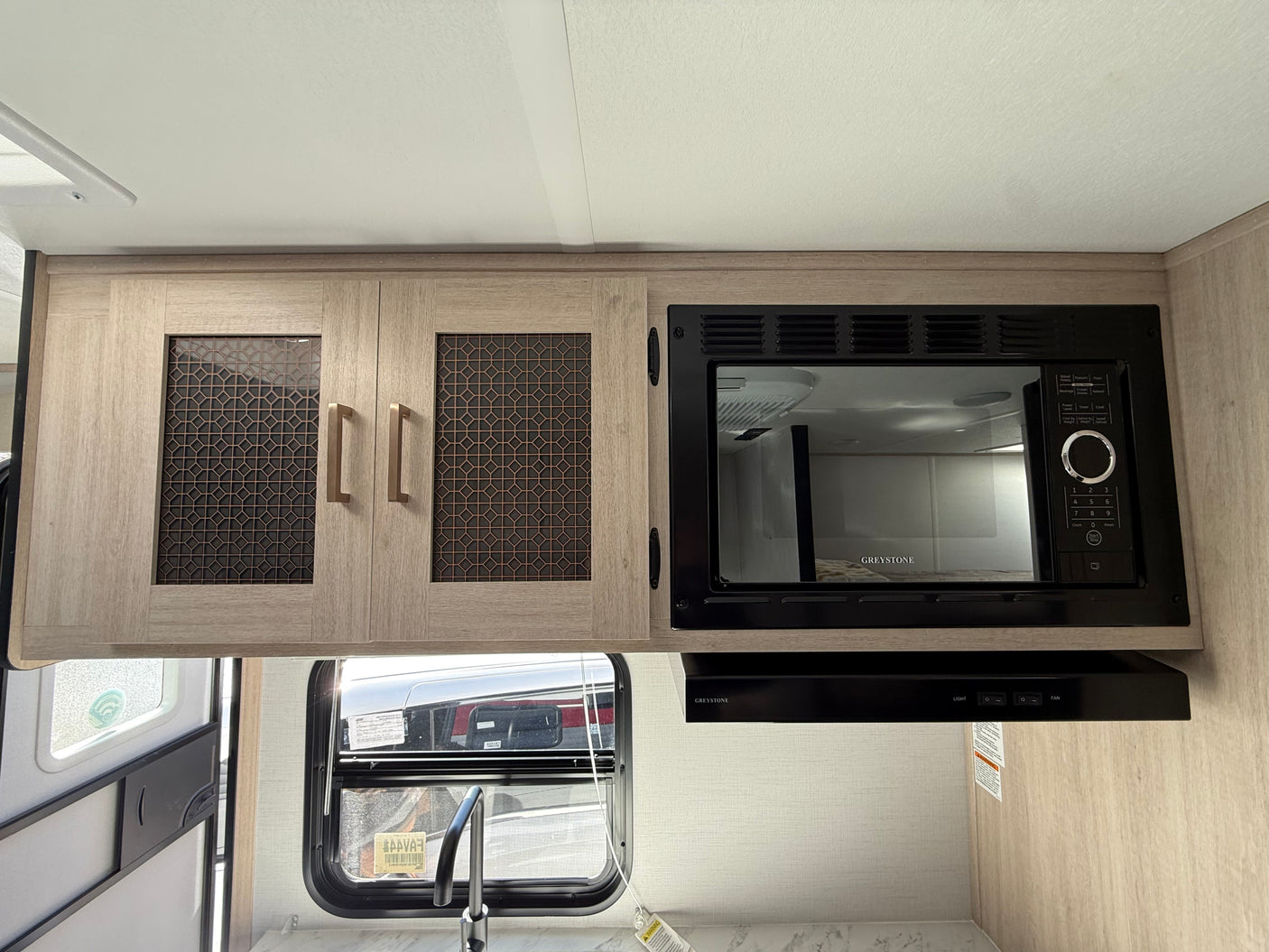 Inside an RV rental kitchen: light wood cabinets, Greystone microwave, range hood, sink, and window view.