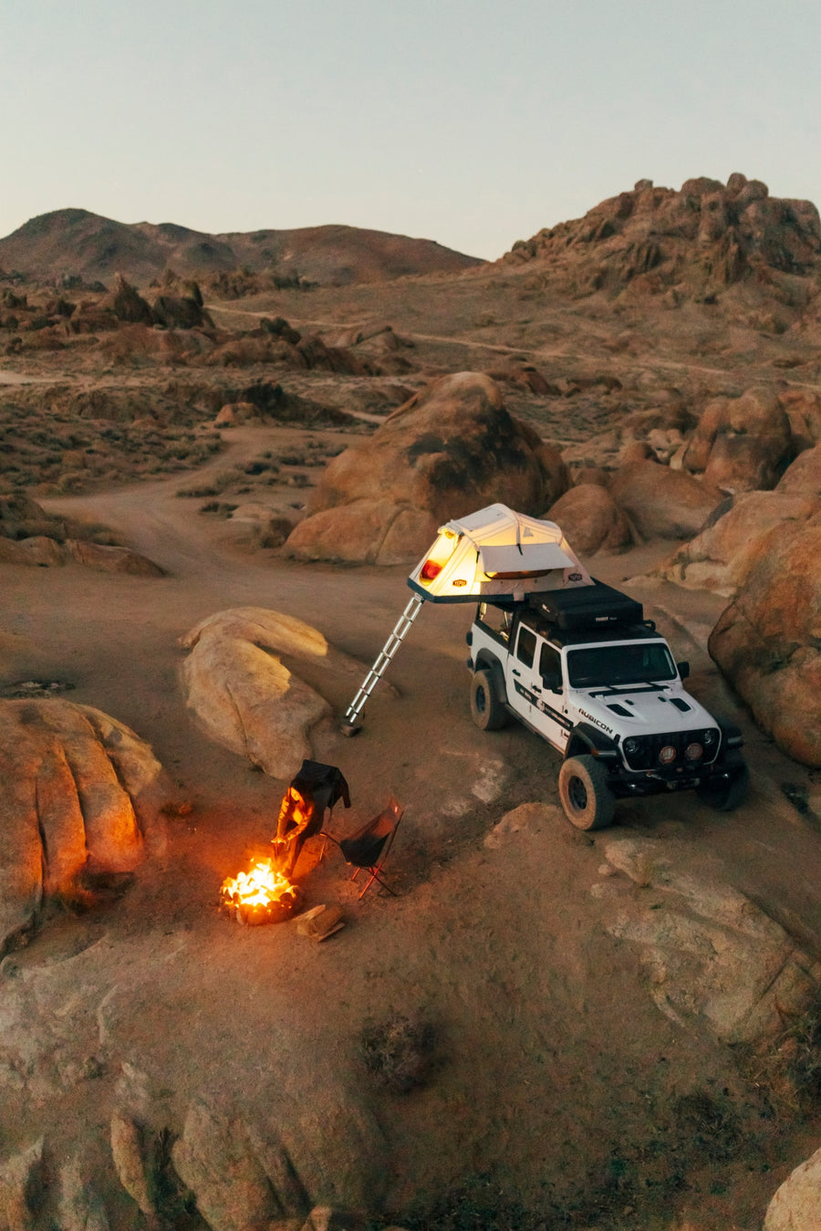 featuring a lit rooftop tent on a white Jeep.|A man sets up a Tepui roof-top tent on a white Jeep 4x4 rv rental for an off-road camping adventure.|Woman organizing gear in her truck's overlanding system for desert camping. Explore outdoor adventures with an RV rental!|Man in a puffer jacket cooking by his overlanding vehicle with a rooftop tent