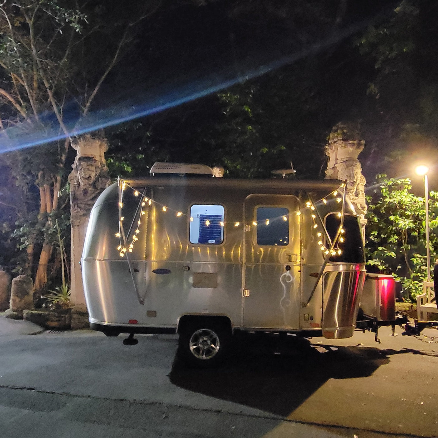 A shiny Airstream RV rental, decorated with star lights, parked at night under a bright lamp near trees.
