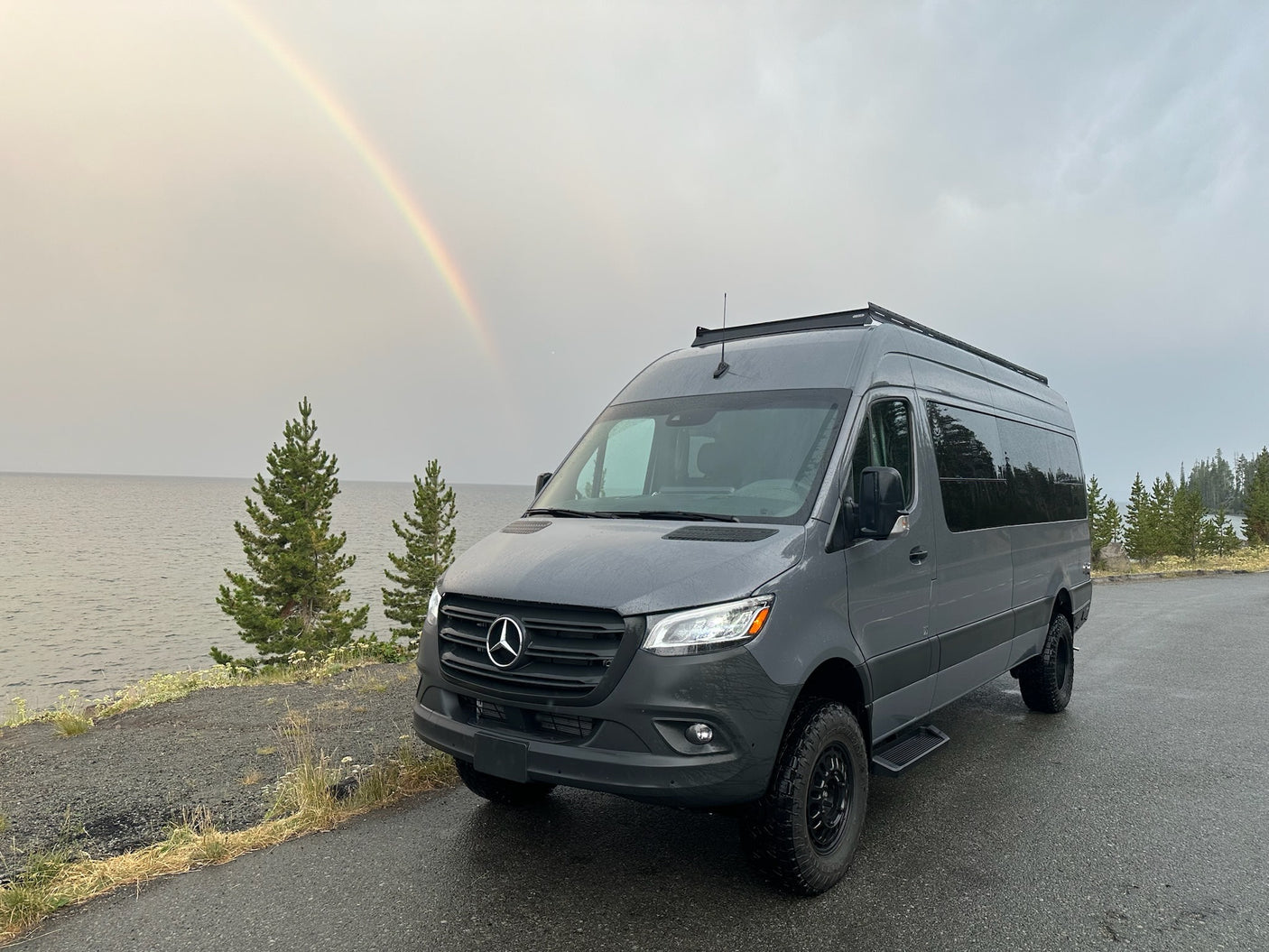 Grey Mercedes-Benz Sprinter **RV rental** parked by a lake. A vibrant rainbow arcs across the cloudy sky.|A gray RV rental on a road