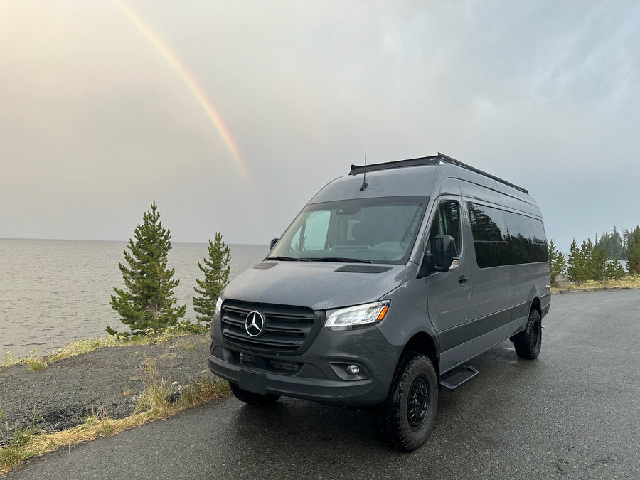 Grey Mercedes-Benz Sprinter **RV rental** parked by a lake. A vibrant rainbow arcs across the cloudy sky.|A gray RV rental on a road