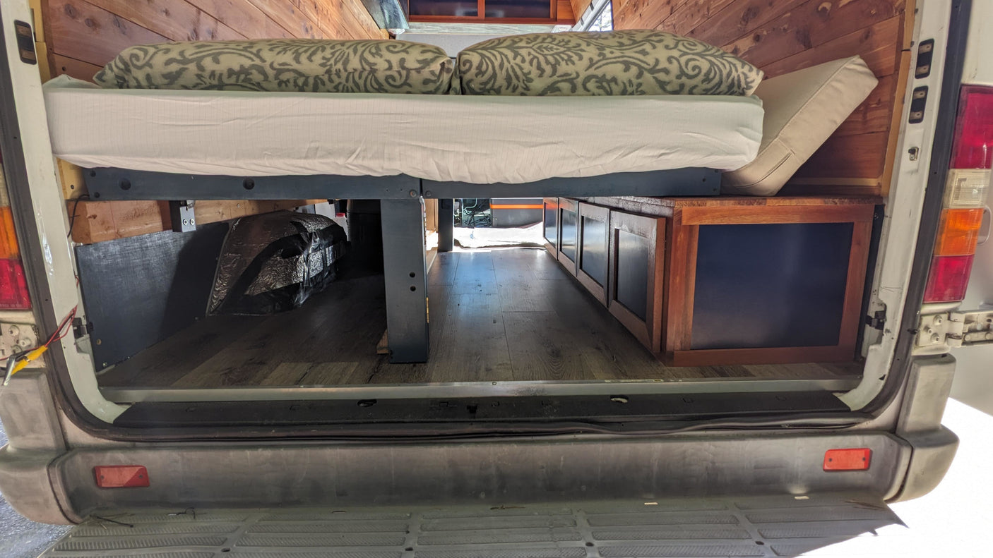 Cozy rv rental camper van interior: raised bed with pillows, under-bed storage cabinets, wood-paneled walls, and laminate flooring.