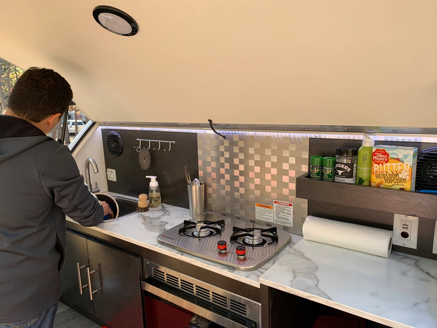 Man washes dishes in a compact RV rental kitchen with a stove and marble-look counters. Ideal for mobile living.