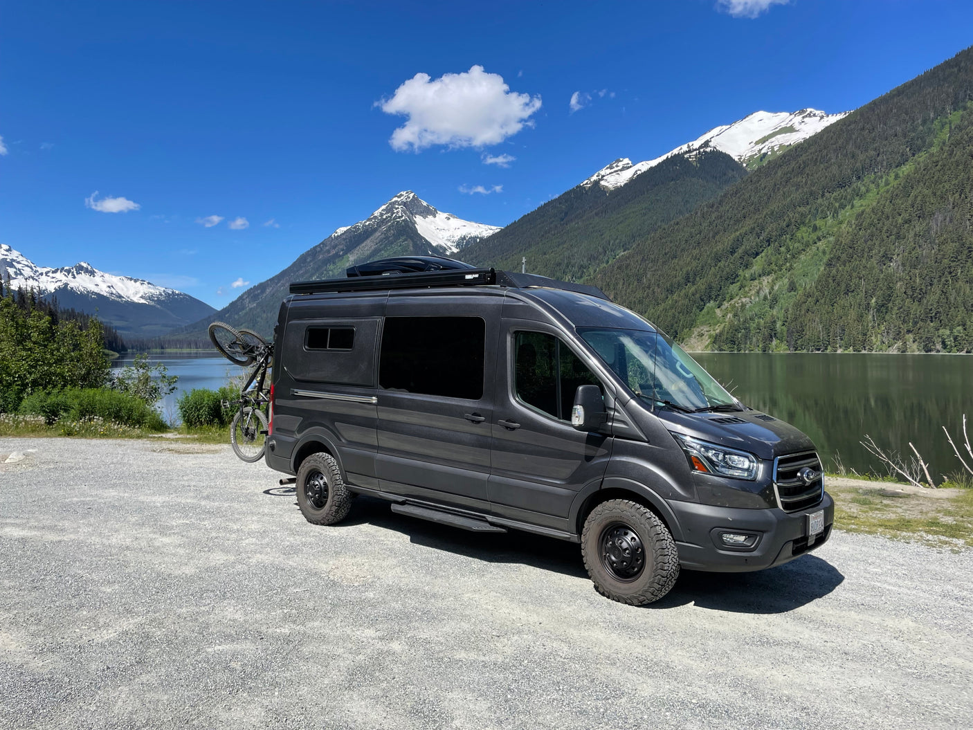 Ford Transit rv rental camper van with bike, parked by a lake amidst majestic snow-capped mountains and lush forests.
