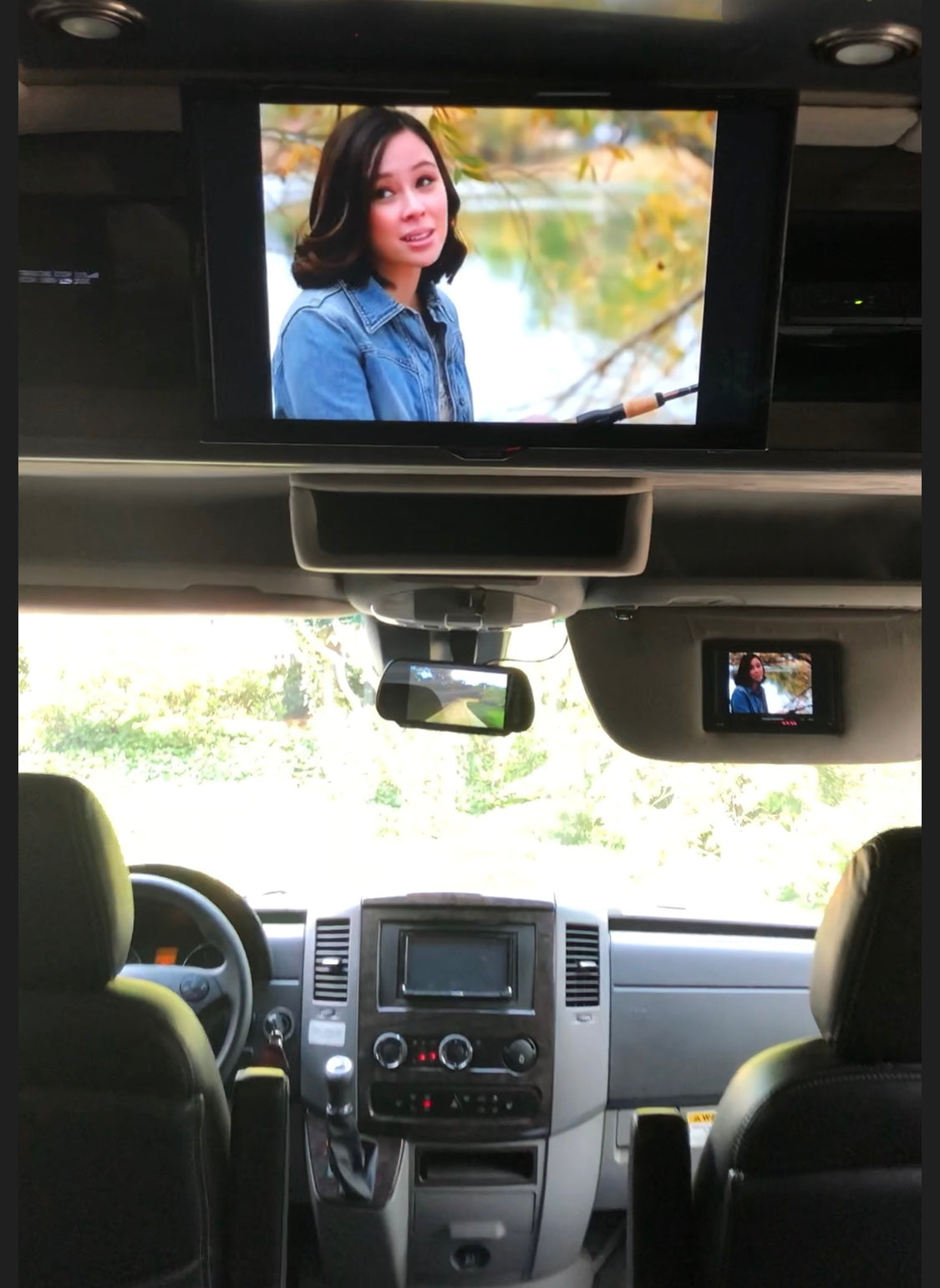 RV rental interior featuring a large overhead screen & a small sun visor screen, both displaying a woman, plus the dashboard.